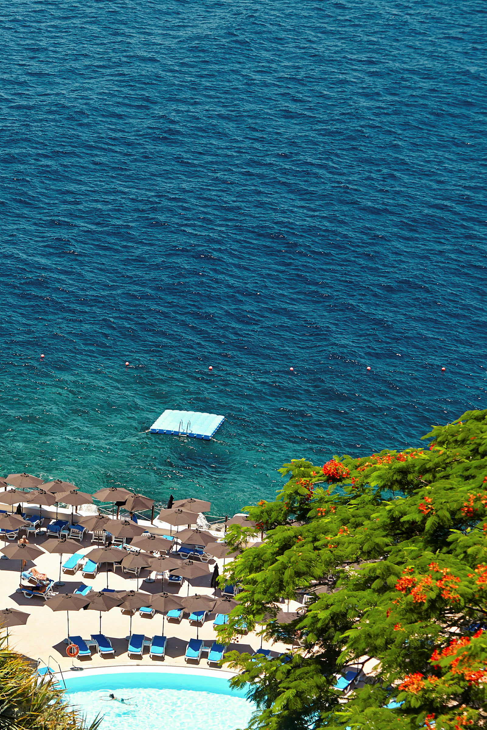 Vy över en strand med parasoller, solstolar, en pool och havet med en flytande plattform. (AI-genererad text)