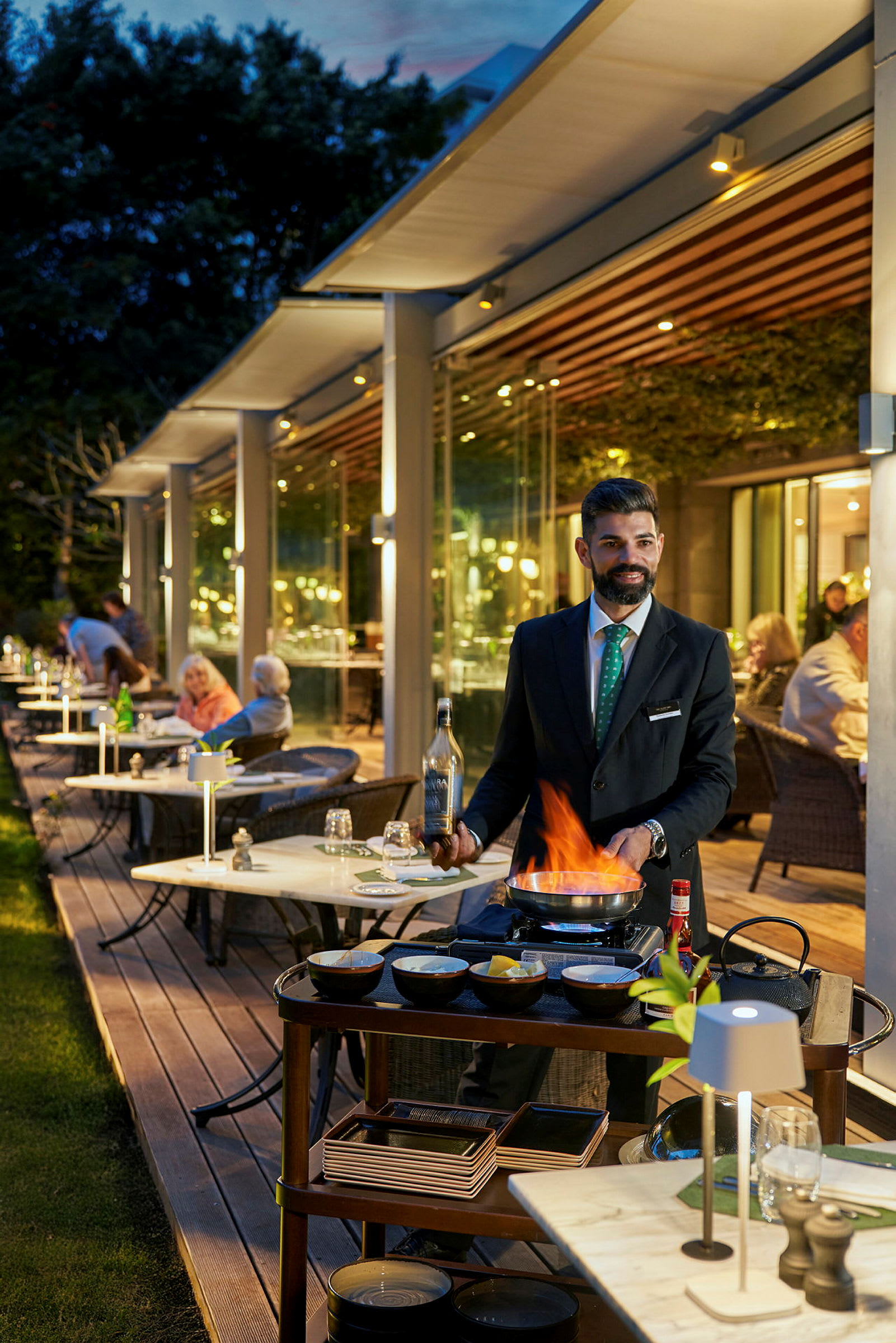En servitör tillagar mat vid ett bord med hjälp av en flamberingsbrännare på en restaurang med uteservering. (AI-genererad text)