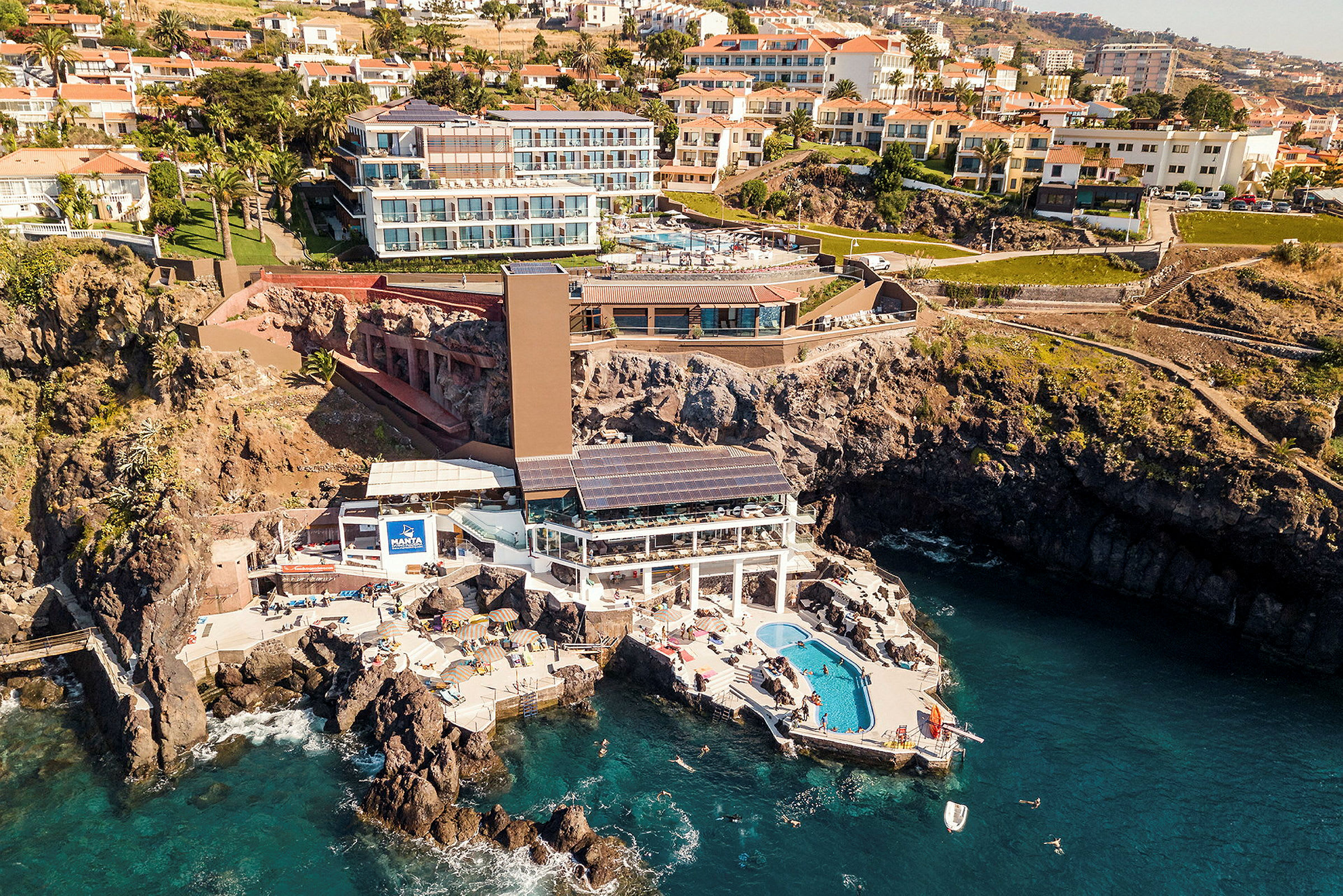 Flygfoto av ett hotell beläget på klippor vid havet, med pool och solstolar. (AI-genererad text)