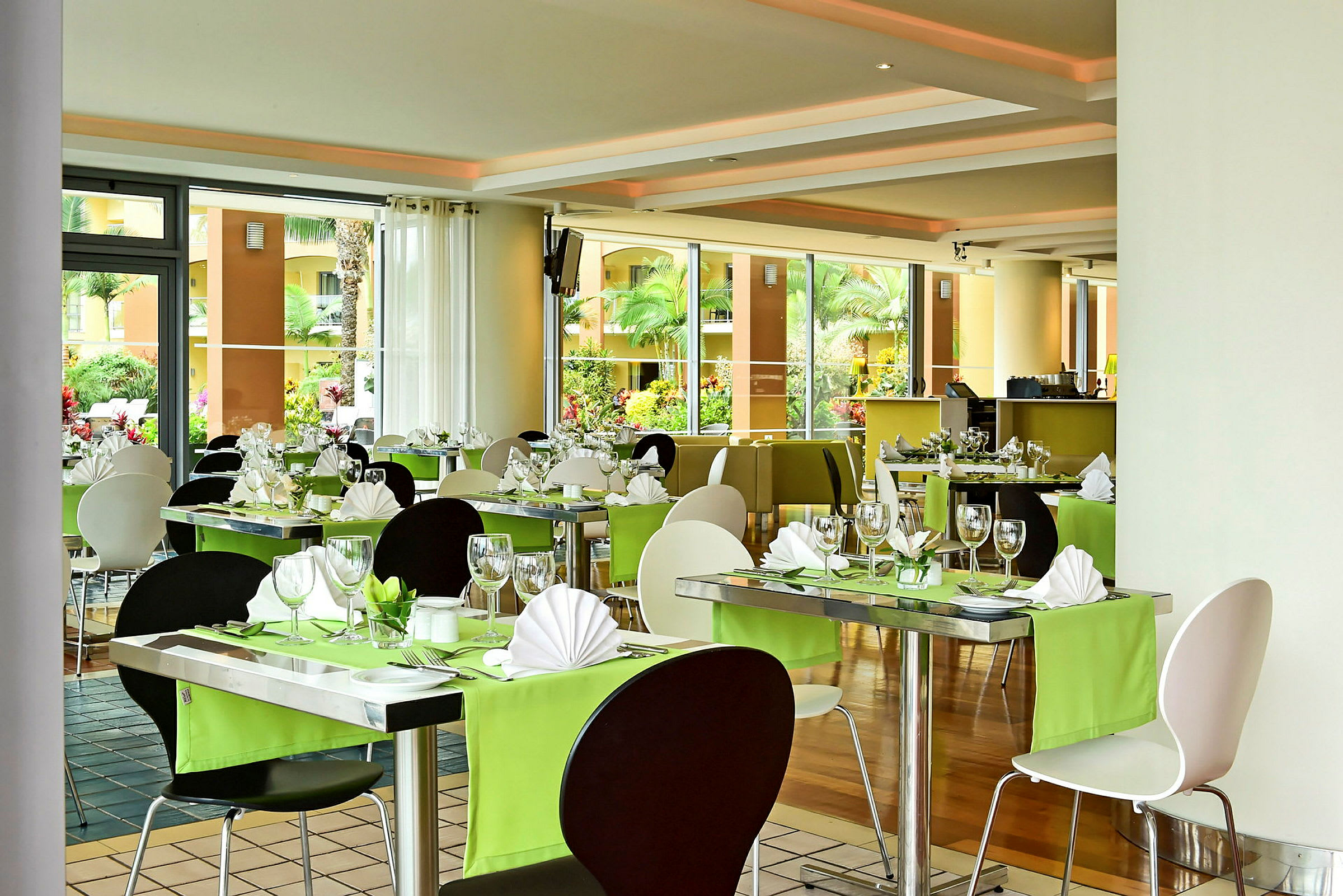 En restaurang med dukade bord, gröna dukar och stora fönster med utsikt över en grön trädgård. (AI-genererad text)