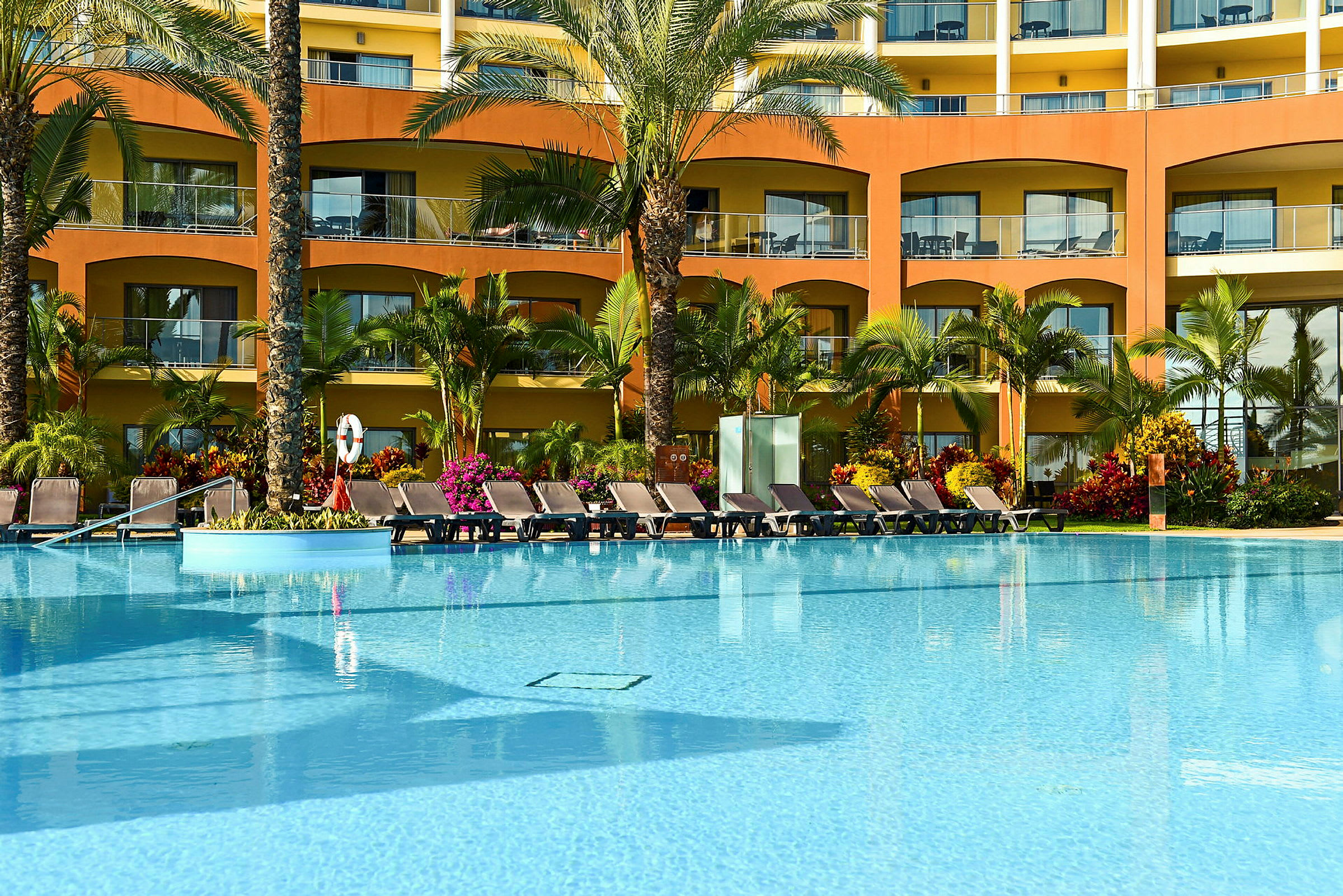 En pool framför ett hotell med solstolar och växter. (AI-genererad text)
