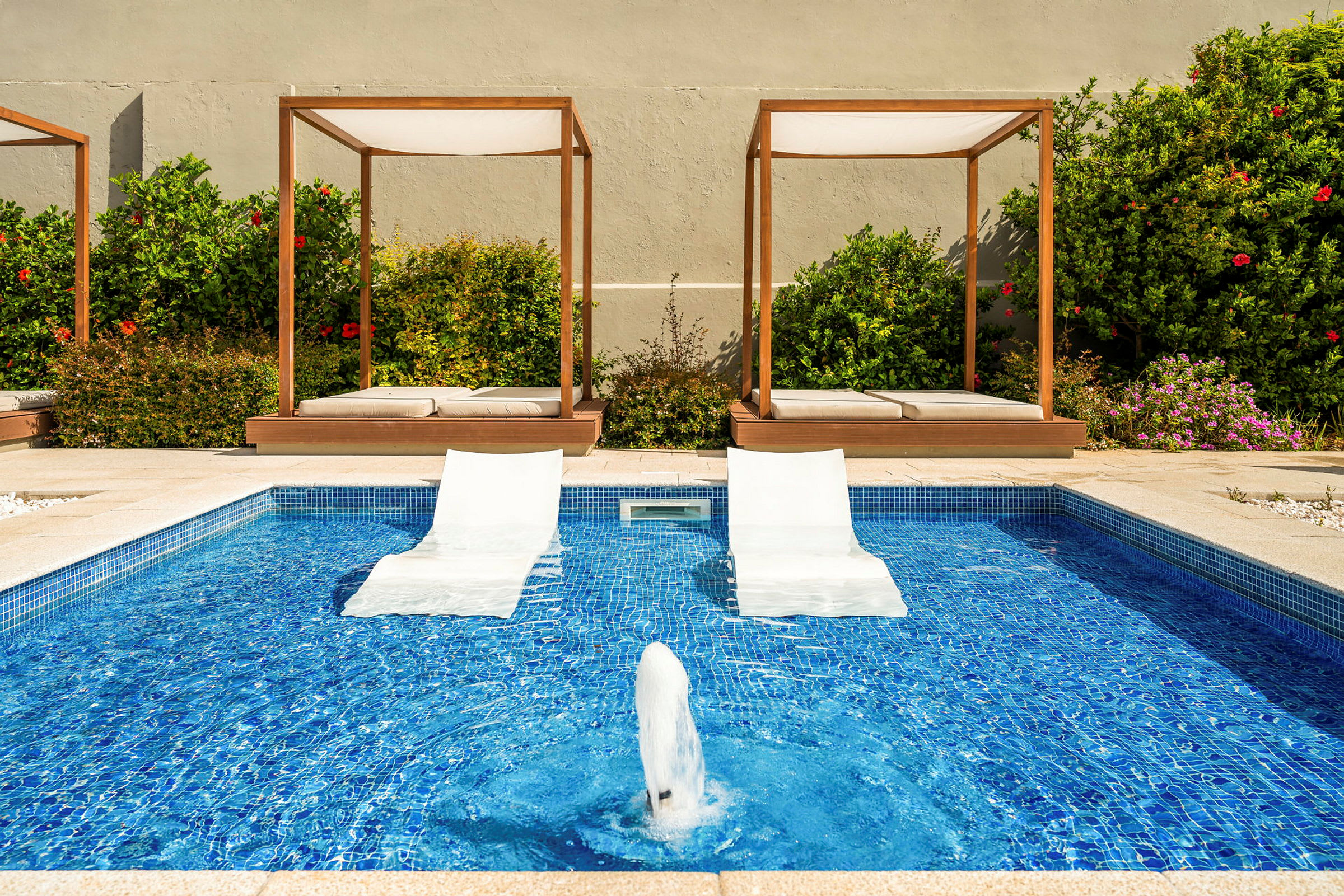 Pool med solstolar i vattnet och solsängar under skuggande struktur framför växtlighet. (AI-genererad text)