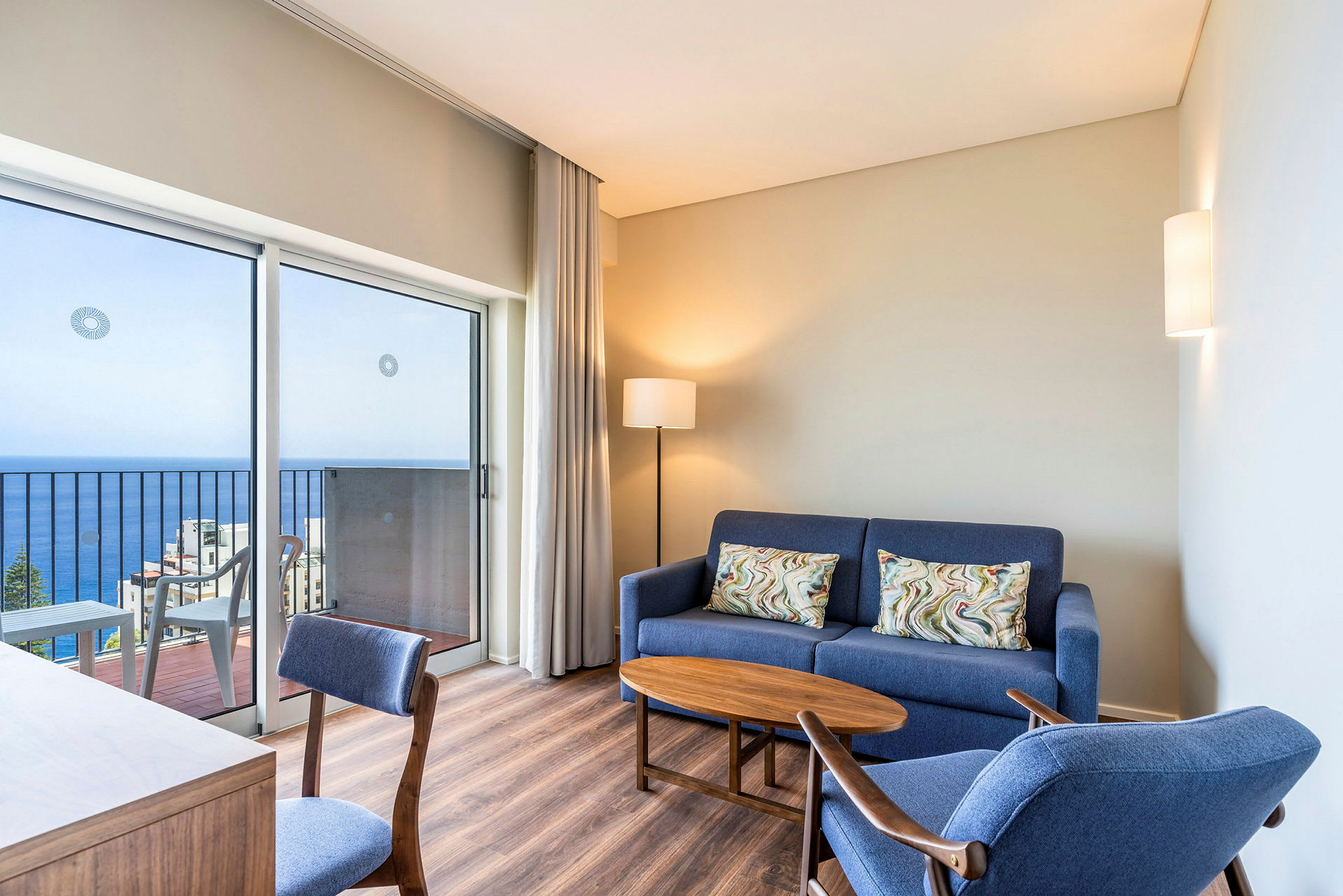Ett modernt inrett hotellrum med en blå soffa, blå sittstol, ett träbord och en skrivbordsstol. Rummet har en balkong med havsutsikt. (AI-genererad text)
