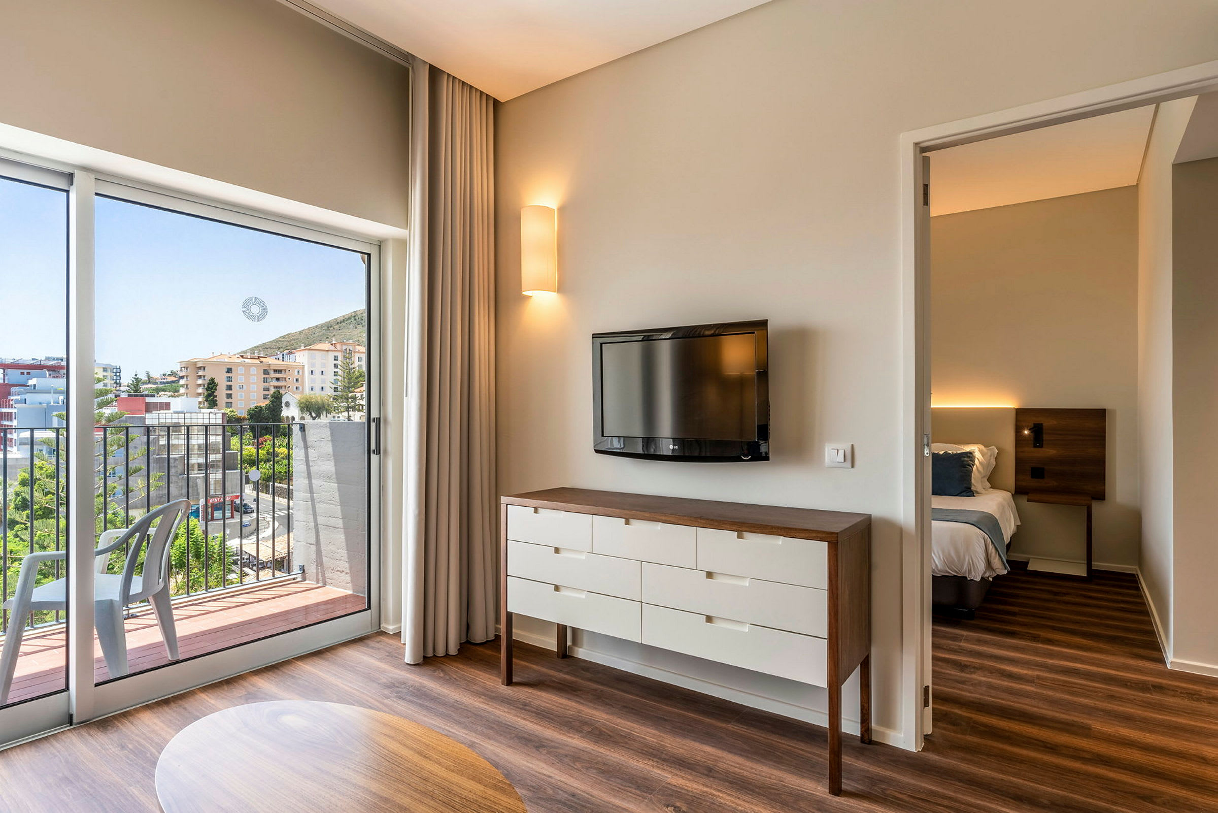 Ett hotellrum med en tv på väggen, en byrå och en säng i ett angränsande rum. Balkongen har utsikt över byggnader och grönytor. (AI-genererad text)