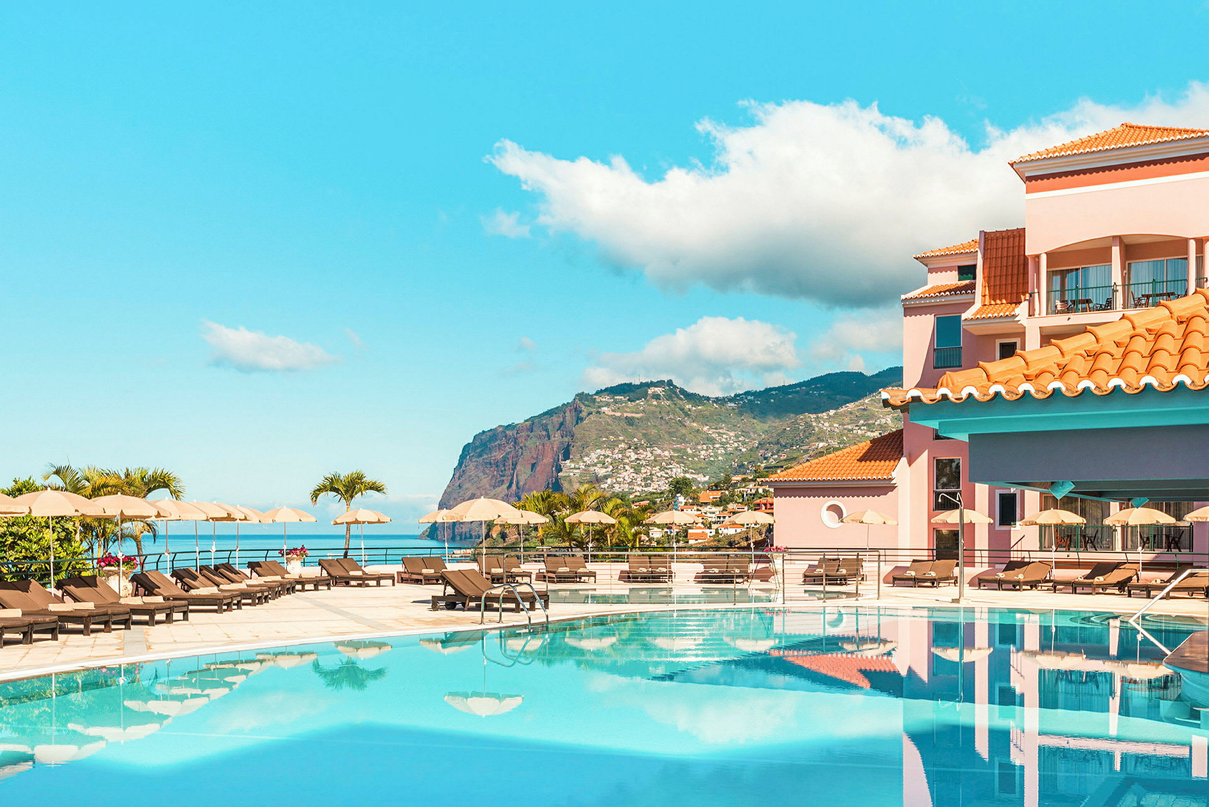 Hotellpool med liggstolar, i bakgrunden utsikt över berg och hav. (AI-genererad text)