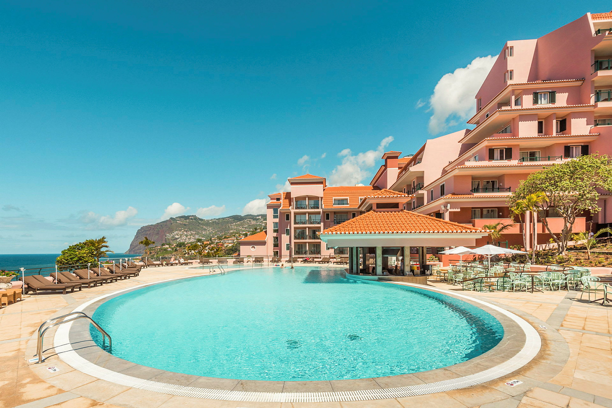En hotellpool omgiven av solstolar och rosa byggnader med havsutsikt. (AI-genererad text)