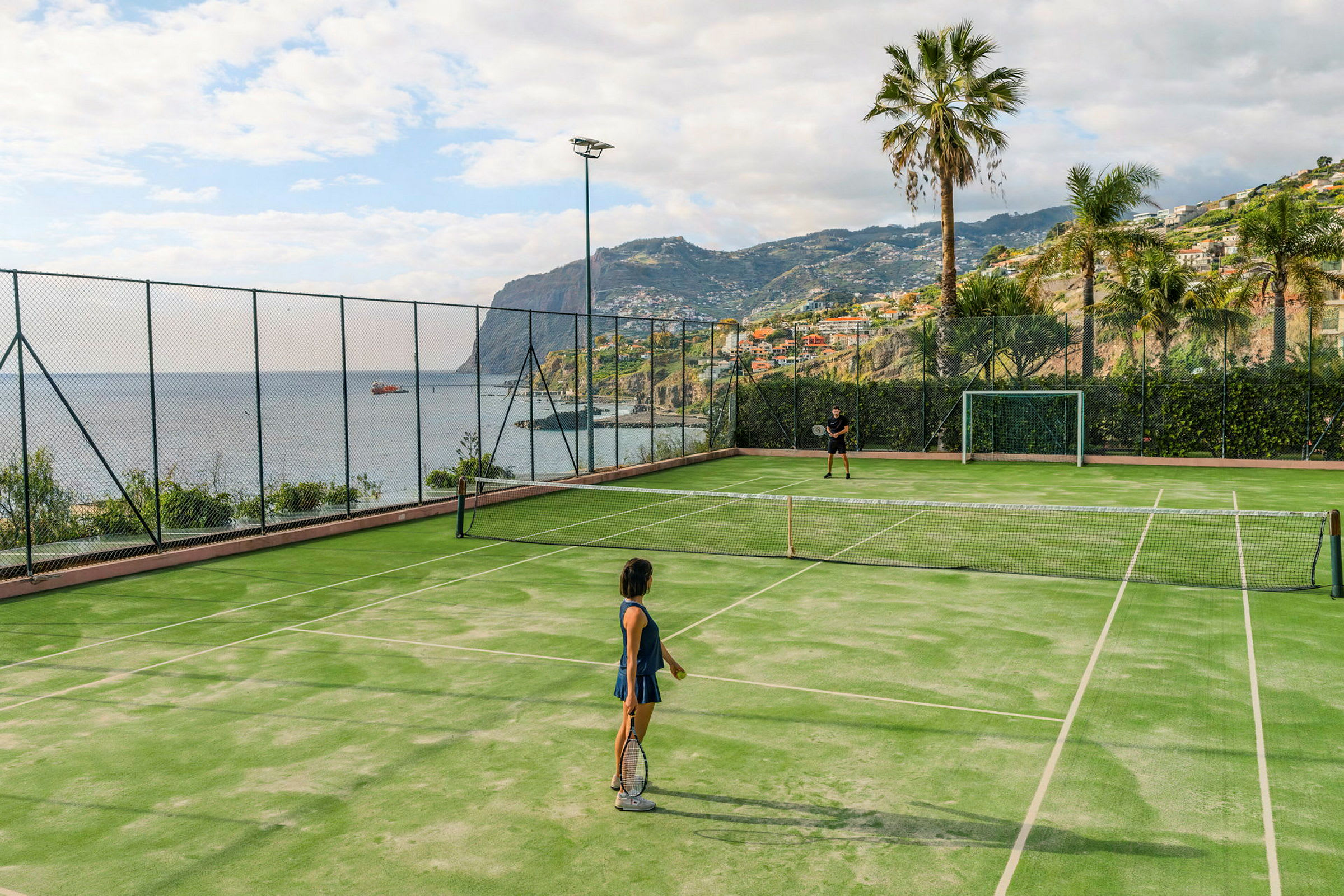 Två personer spelar tennis på en utomhusbana med utsikt över havet och omgivande berg. (AI-genererad text)