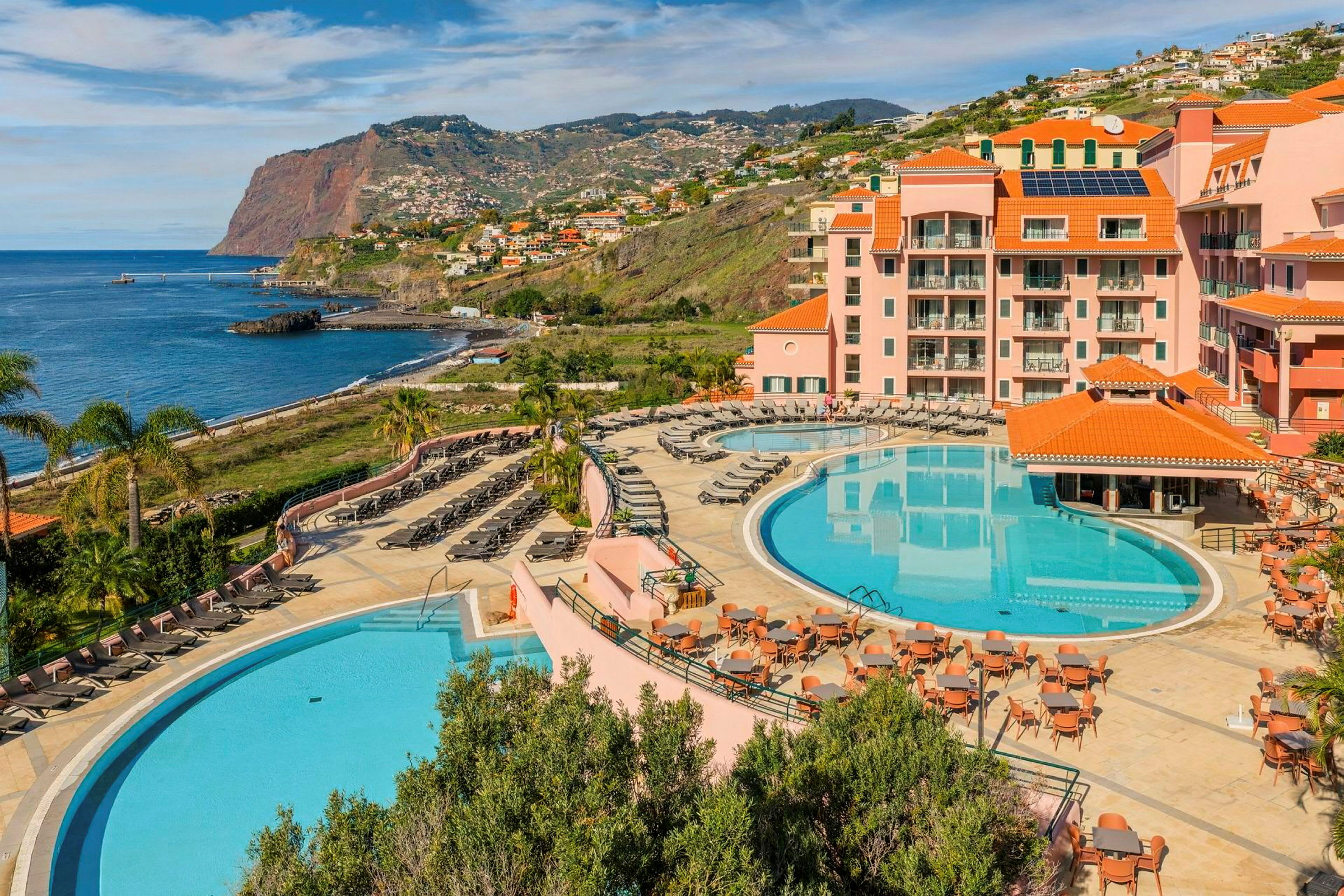 Ett hotellområde vid kusten med två stora pooler, solstolar, bord, och omgivande byggnader med orange tak. Bakgrunden visar en klippa vid havet och bostadsområden längs sluttningen. (AI-genererad text)