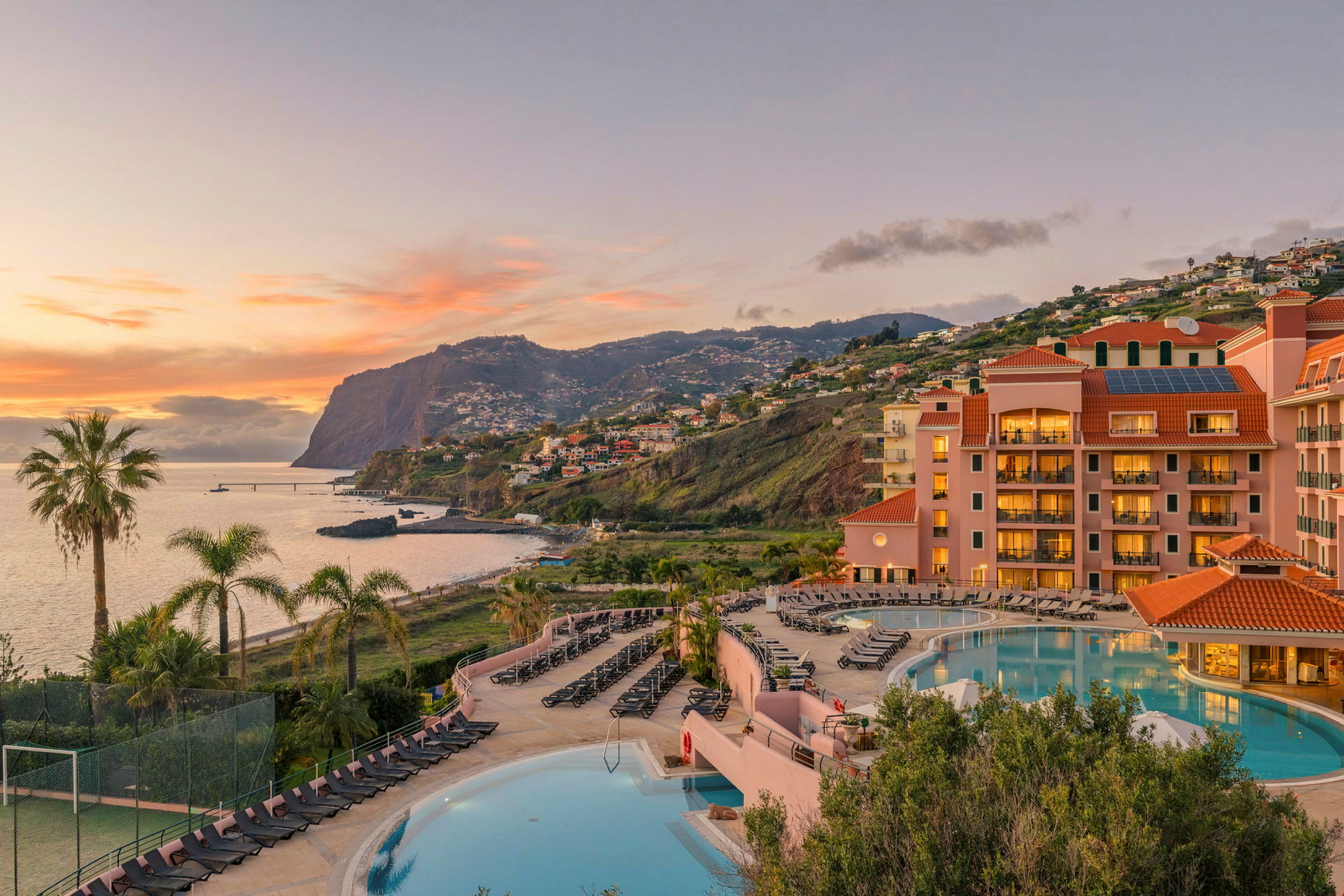 Ett hotell med en swimmingpool i förgrunden och en fantastisk vy över havet och bergen i bakgrunden. (AI-genererad text)