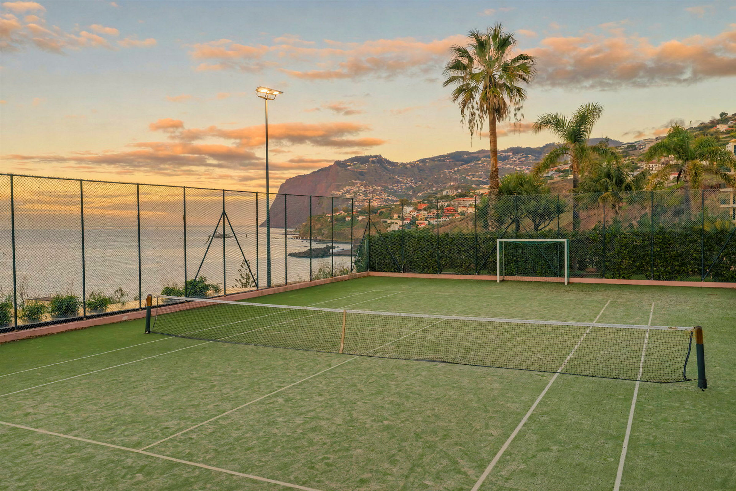 En tennisbana nära havet, omgiven av stängsel, palmer och berg i bakgrunden. (AI-genererad text)