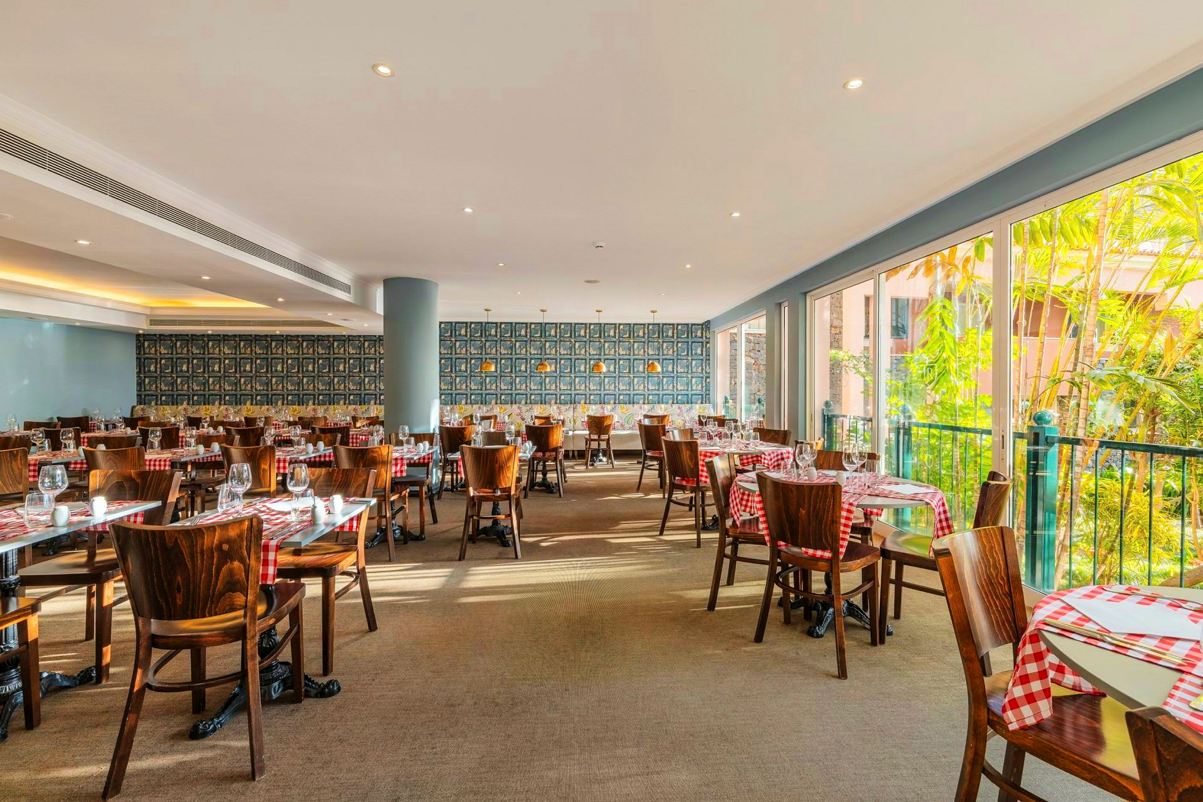 En ljust inredd restauranglokal med rödrutiga dukar på träbord och stolar. Stora fönster släpper in dagsljus och visar gröna växter utanför. (AI-genererad text)