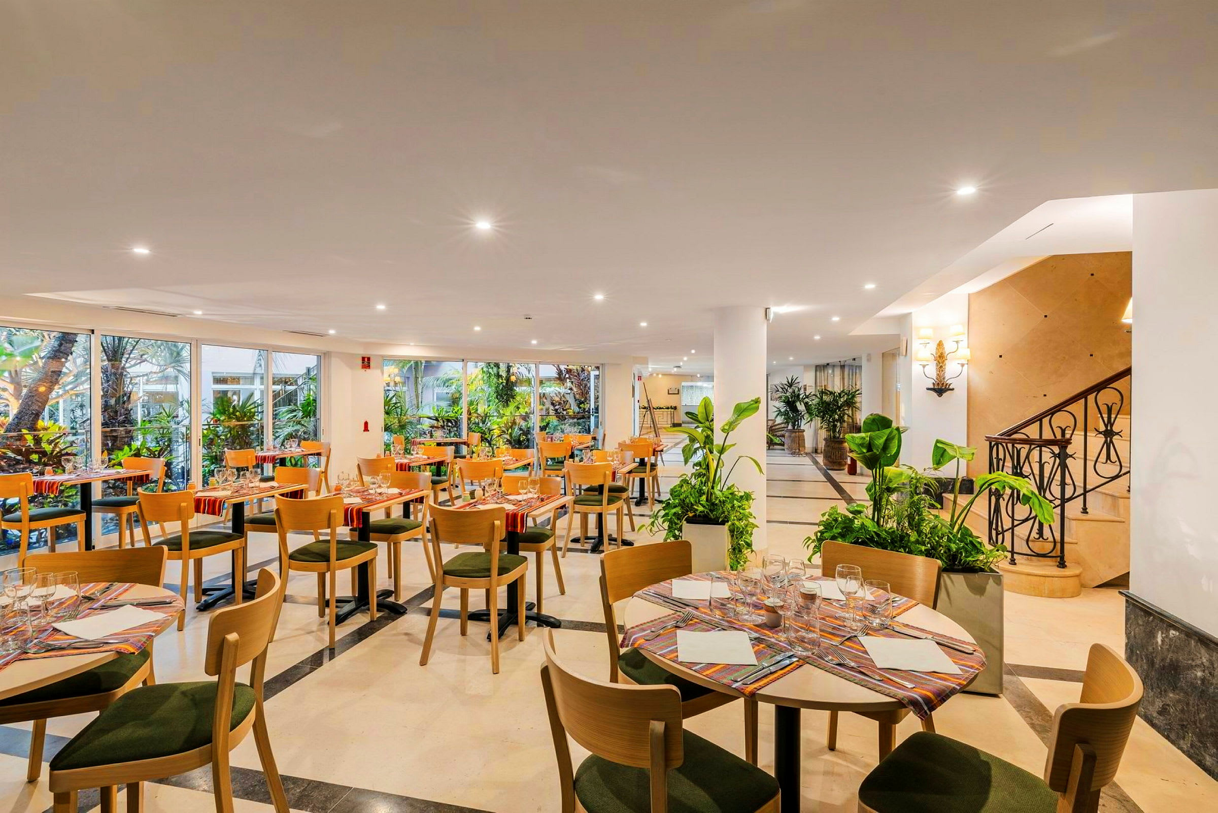En väl upplyst restaurang med flera dukade bord omgiven av glasfönster och växter. (AI-genererad text)