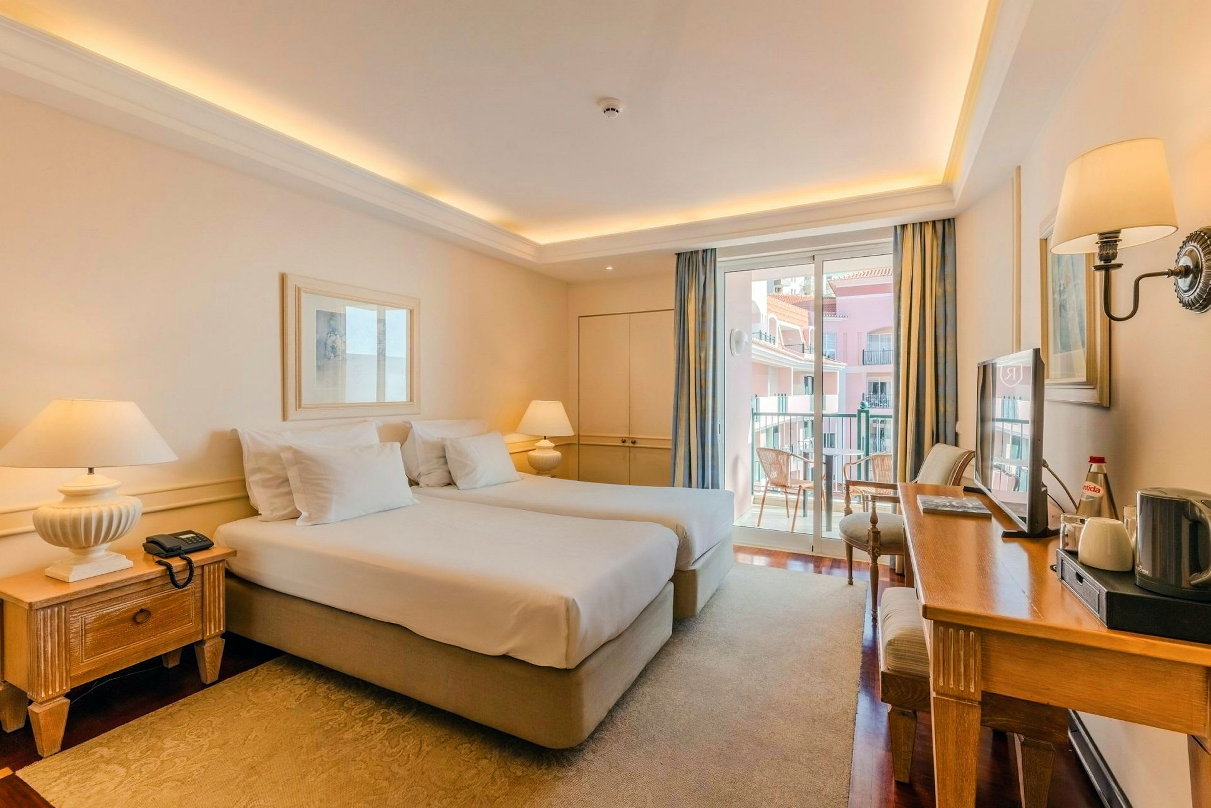 Ett ljust hotellrum med två enkelsängar, ett skrivbord, och en balkong som vetter mot andra byggnader. (AI-genererad text)
