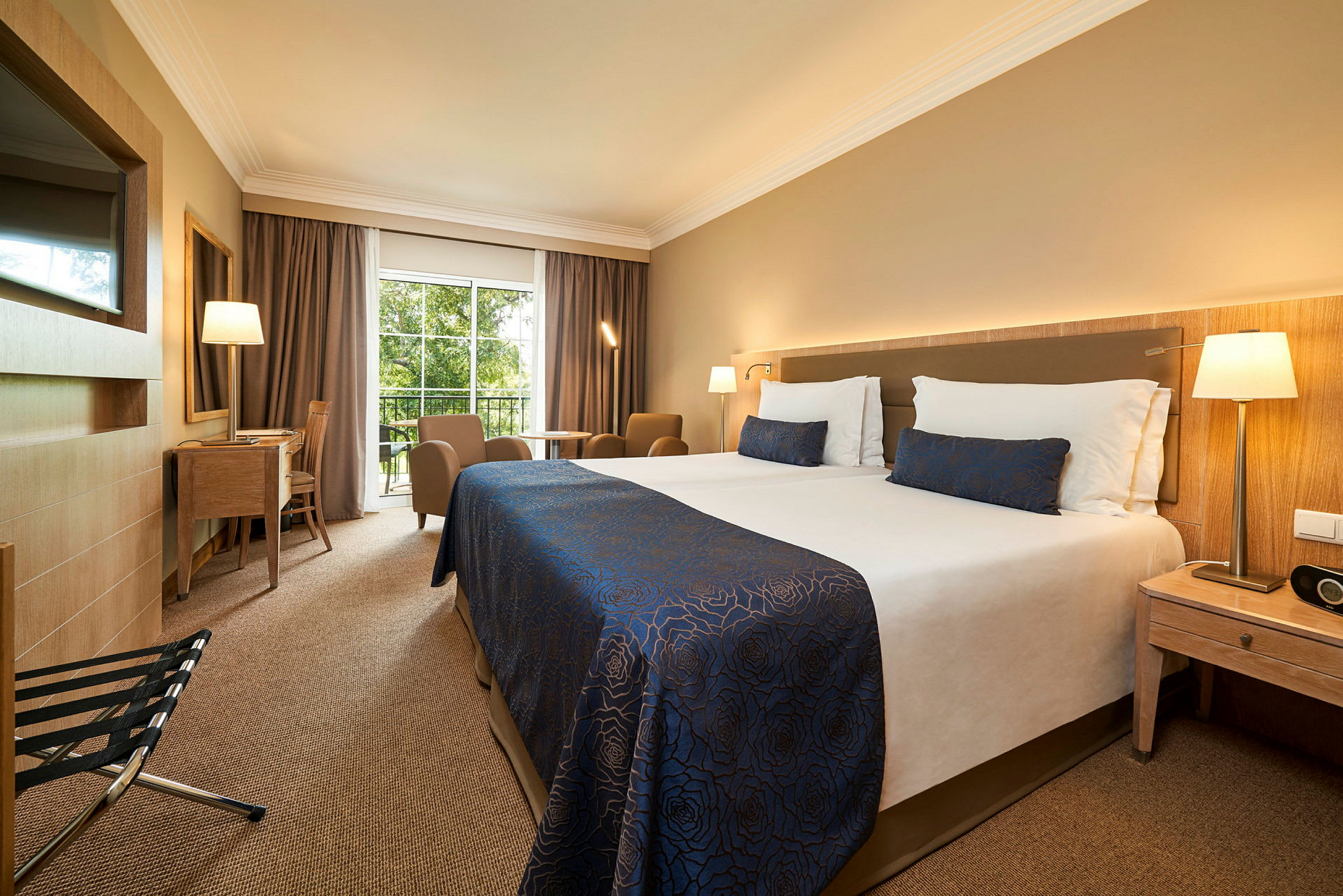 Hotellrum med dubbelsäng, sittområde, skrivbord och utsikt mot grönska. (AI-genererad text)