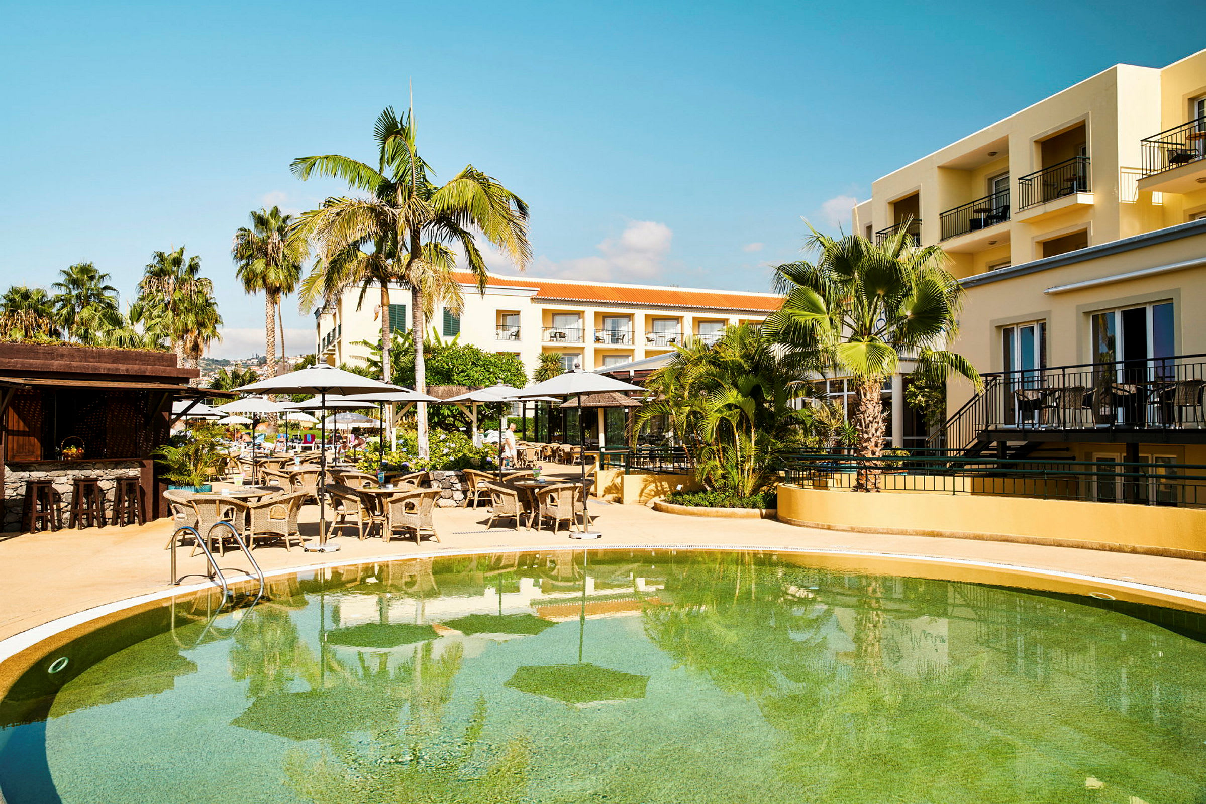 Bild av ett hotellområde med en pool, solstolar och parasoller, omringad av palmer och gulmålad byggnad. (AI-genererad text)