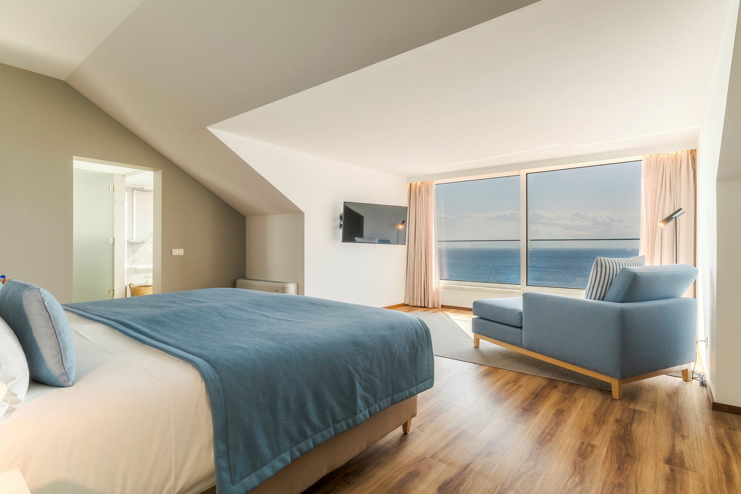 Modernt hotellrum med havsutsikt, säng med blå överkast och blå soffa. (AI-genererad text)