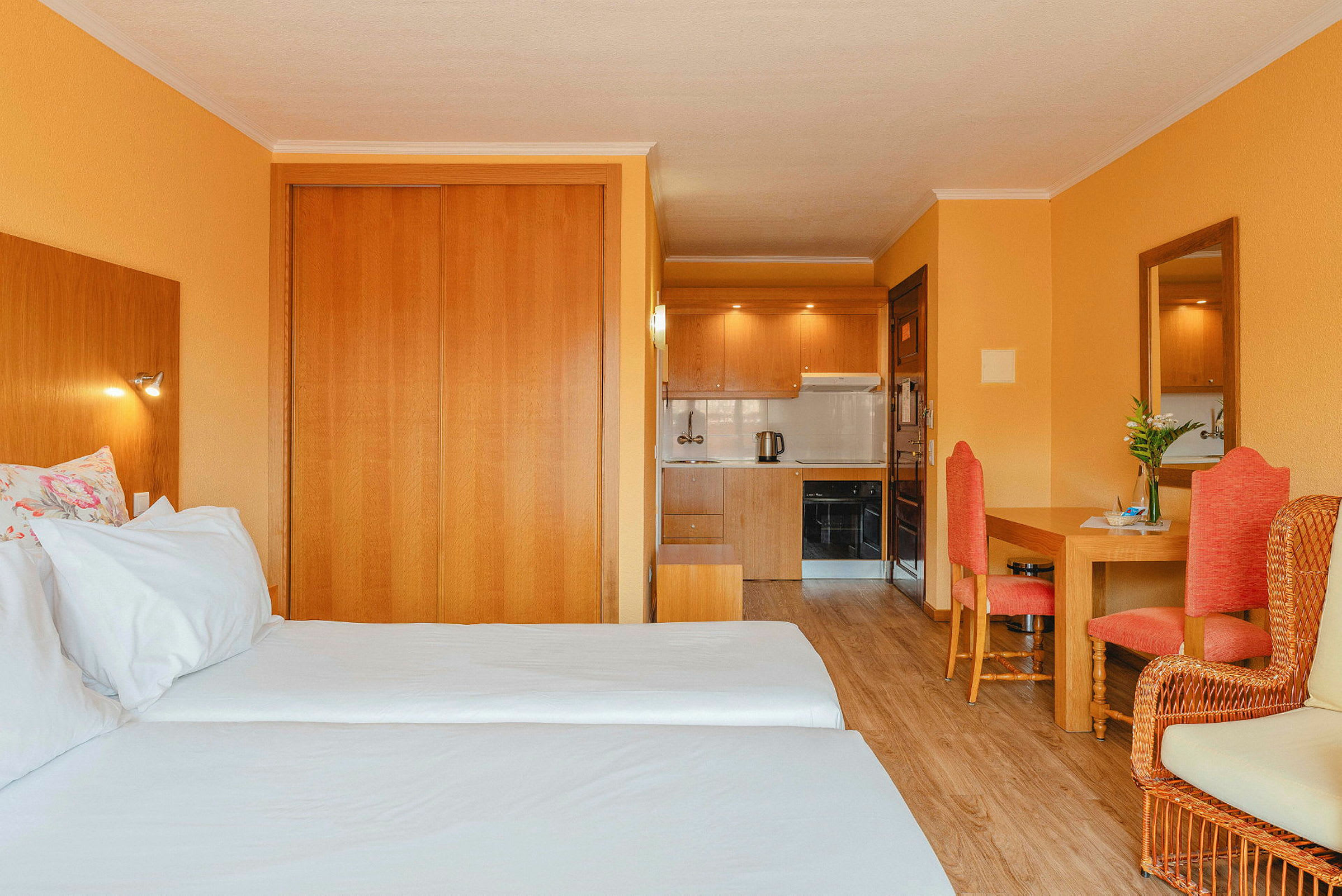 Hotellrum med två enkelsängar, trämöbler, orange väggar och ett litet kök i bakgrunden. (AI-genererad text)