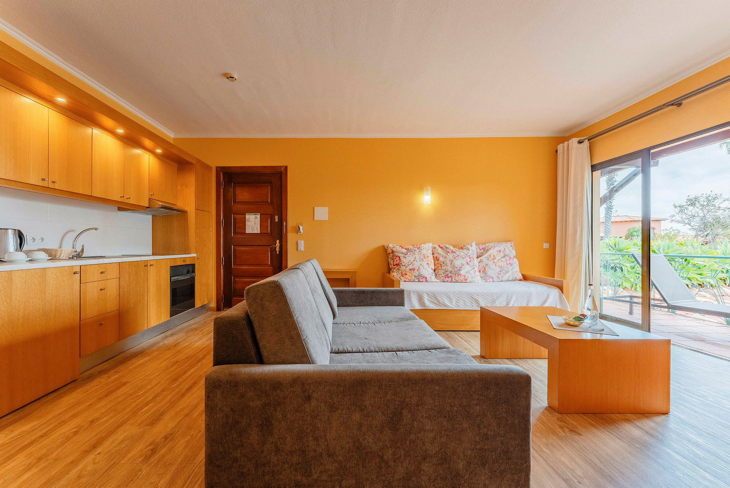 En bild av ett rymligt hotellrum med öppen planlösning. Rummet har ett fullt utrustat kök med träskåp, en sittgrupp med en grå soffa, ett soffbord och en bädd med färgglada kuddar. Glasdörrar leder ut till en terrass med utsikt över grönska. (AI-genererad text)
