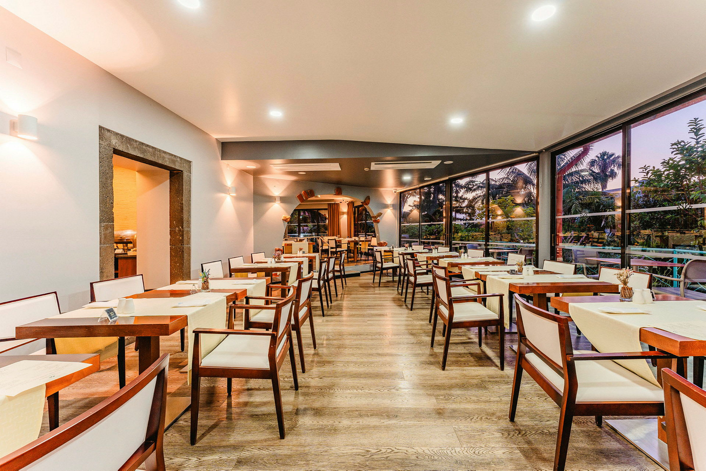 Bild av en restaurang med tomma bord och stolar, träinredning och stora fönster som vetter mot en trädgård. (AI-genererad text)