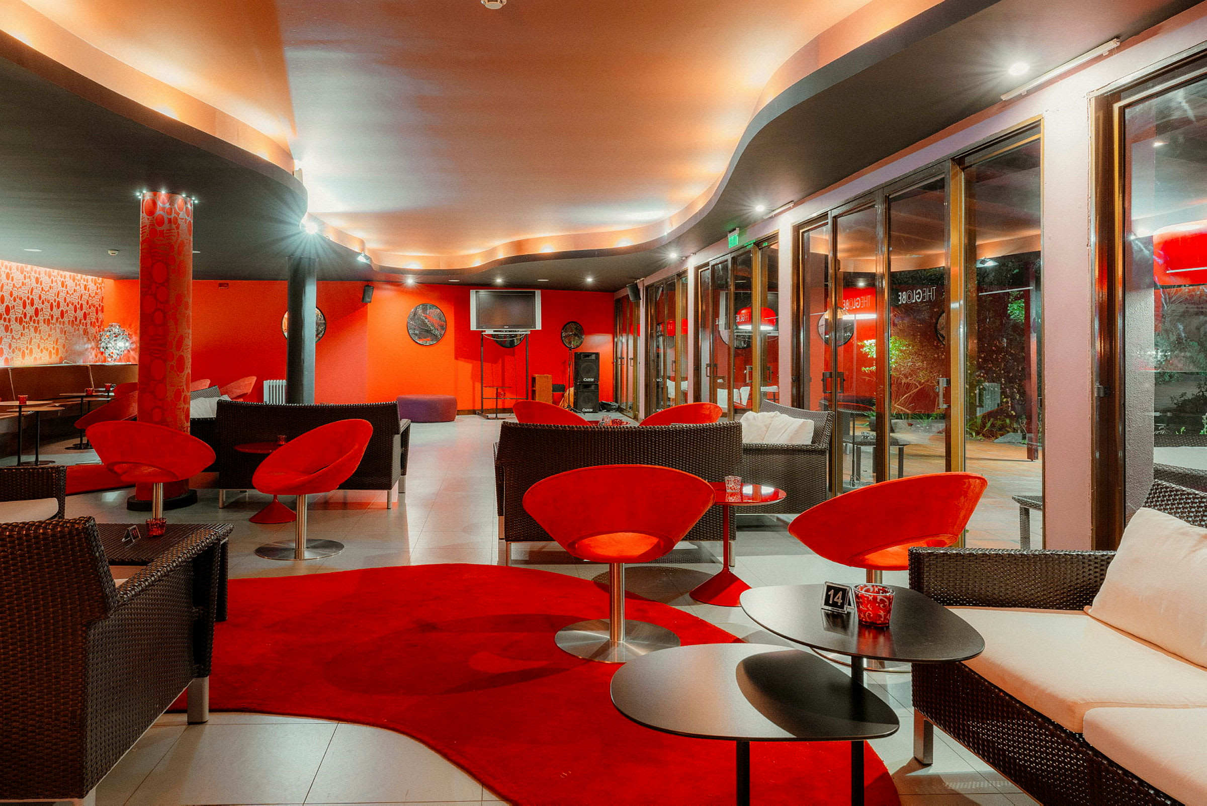 Interiör av en smakfullt inredd lounge med röda och mörka möbler, mjuk belysning och stora fönster. (AI-genererad text)