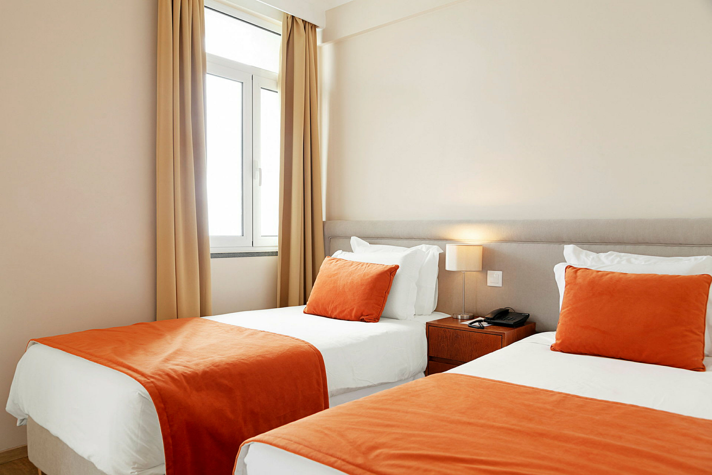 Ett hotellrum med två enkelsängar bredvid varandra, med vita lakan och orange överkast och prydnadskuddar. (AI-genererad text)