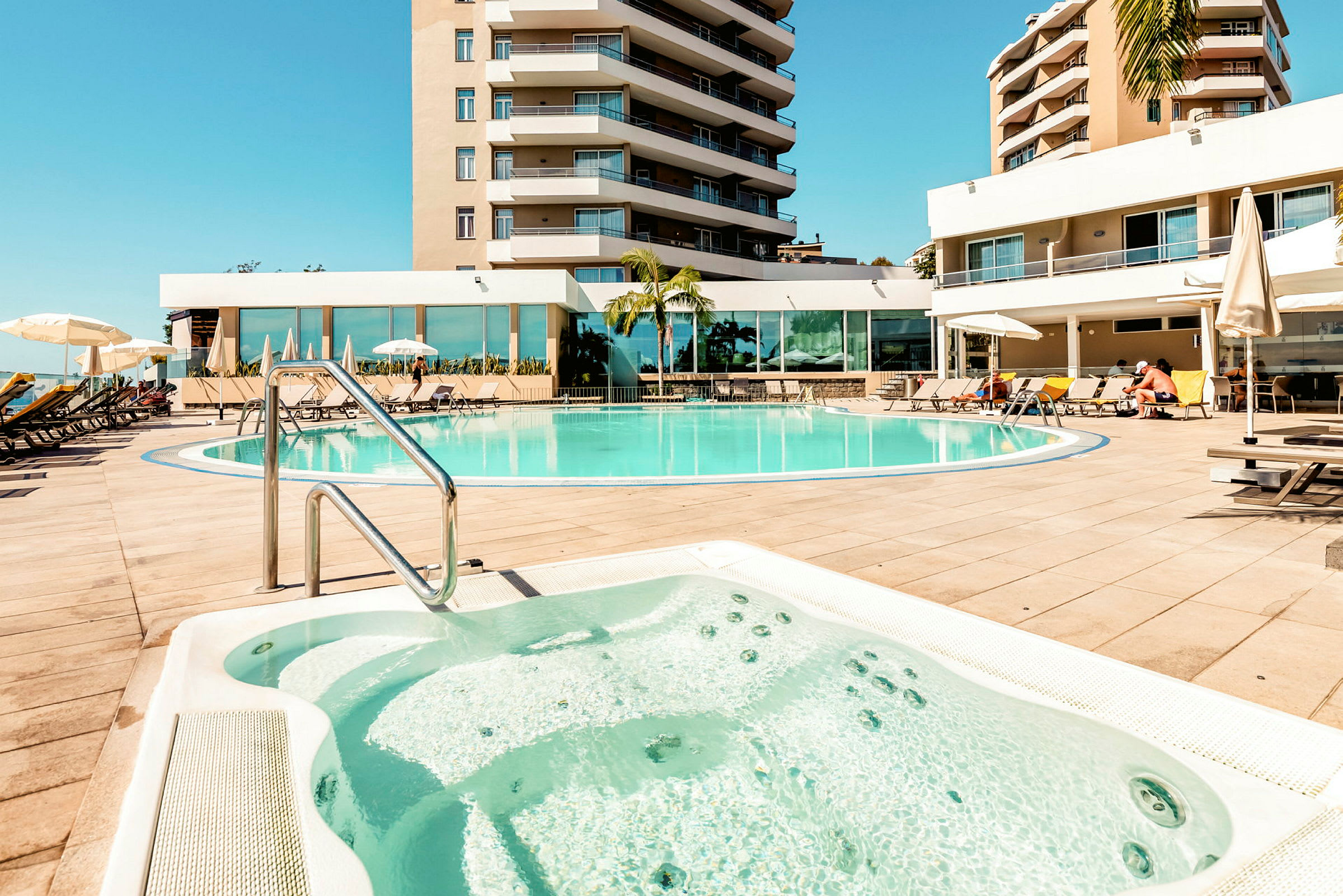 Bild av ett hotellpoolområde med en jacuzzi i förgrunden, solstolar och parasoller samt omgivande byggnader. (AI-genererad text)