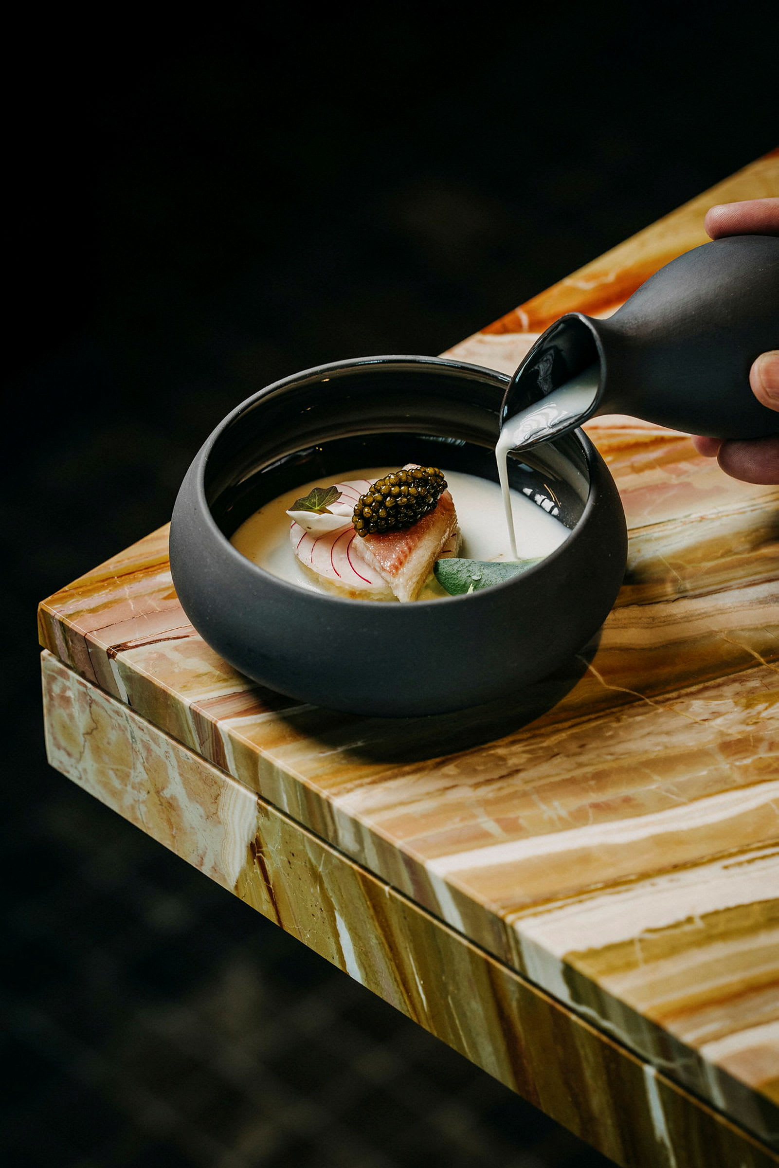 En mörk skål med en maträtt på ett marmorbord. En hand häller vitt sås från en liten kanna. (AI-genererad text)