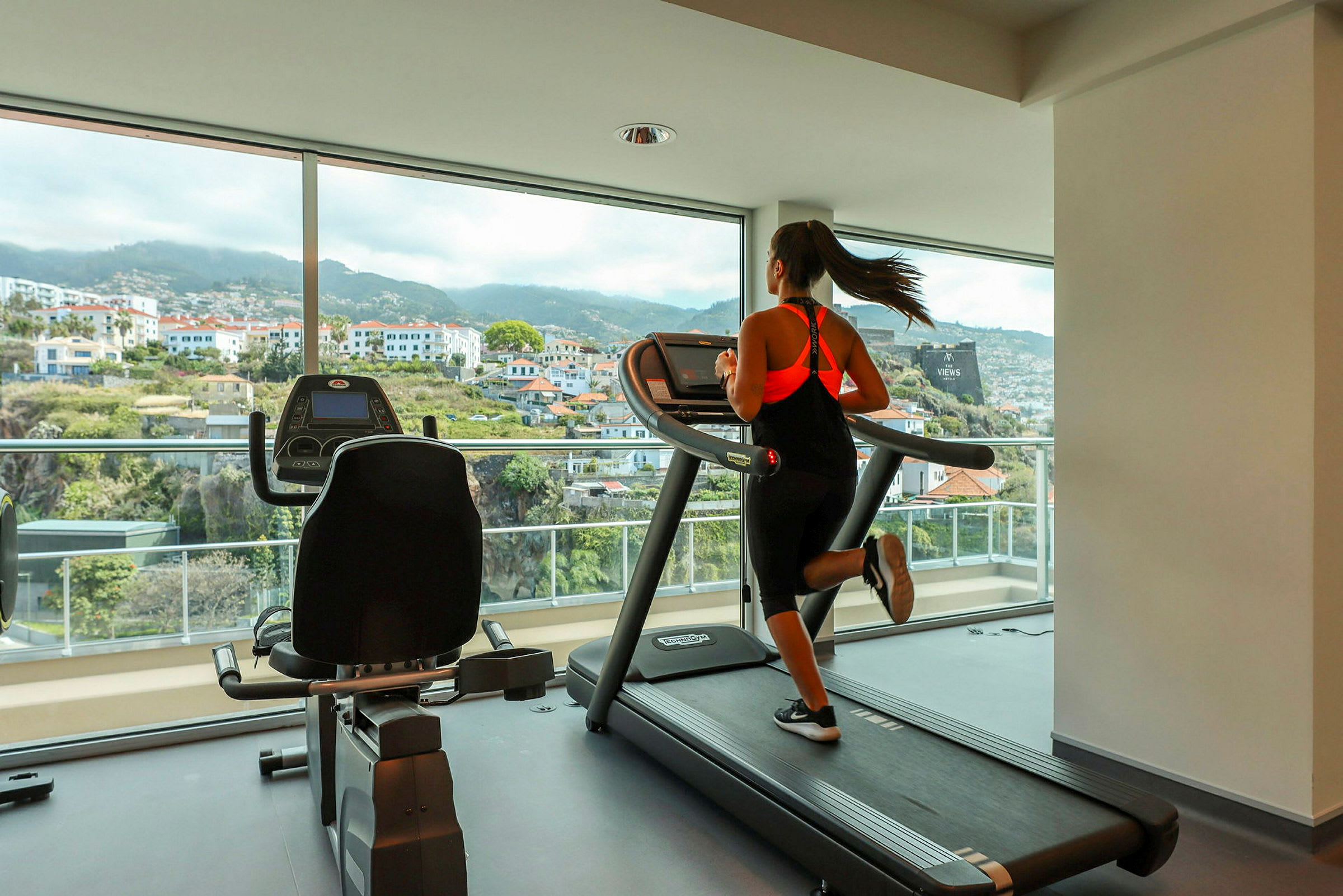 En person springer på löpband i ett gym med stora fönster och utsikt över staden. (AI-genererad text)