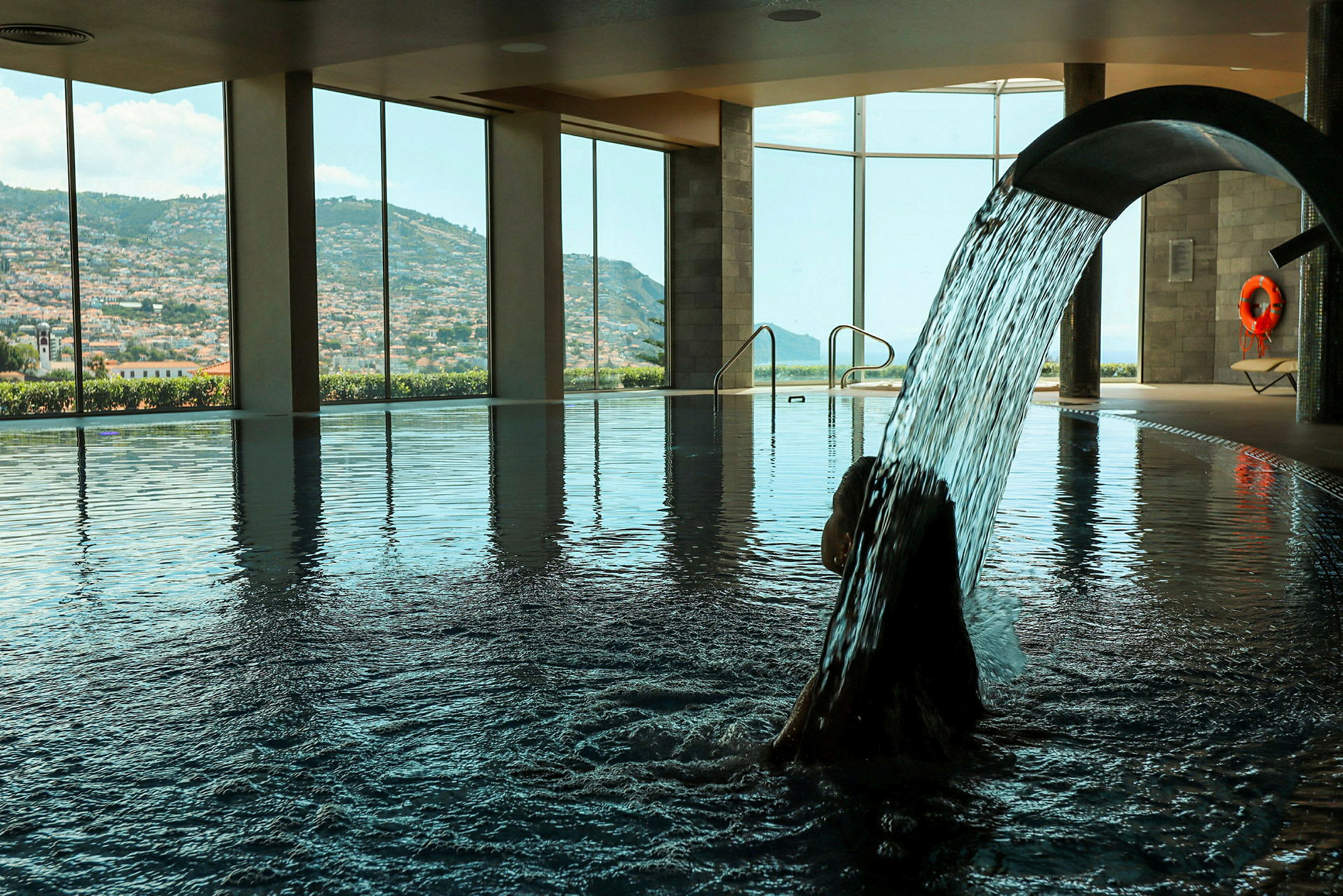 Inomhuspool med panoramautsikt över en bergsstad och en person under en vattenstråle. (AI-genererad text)