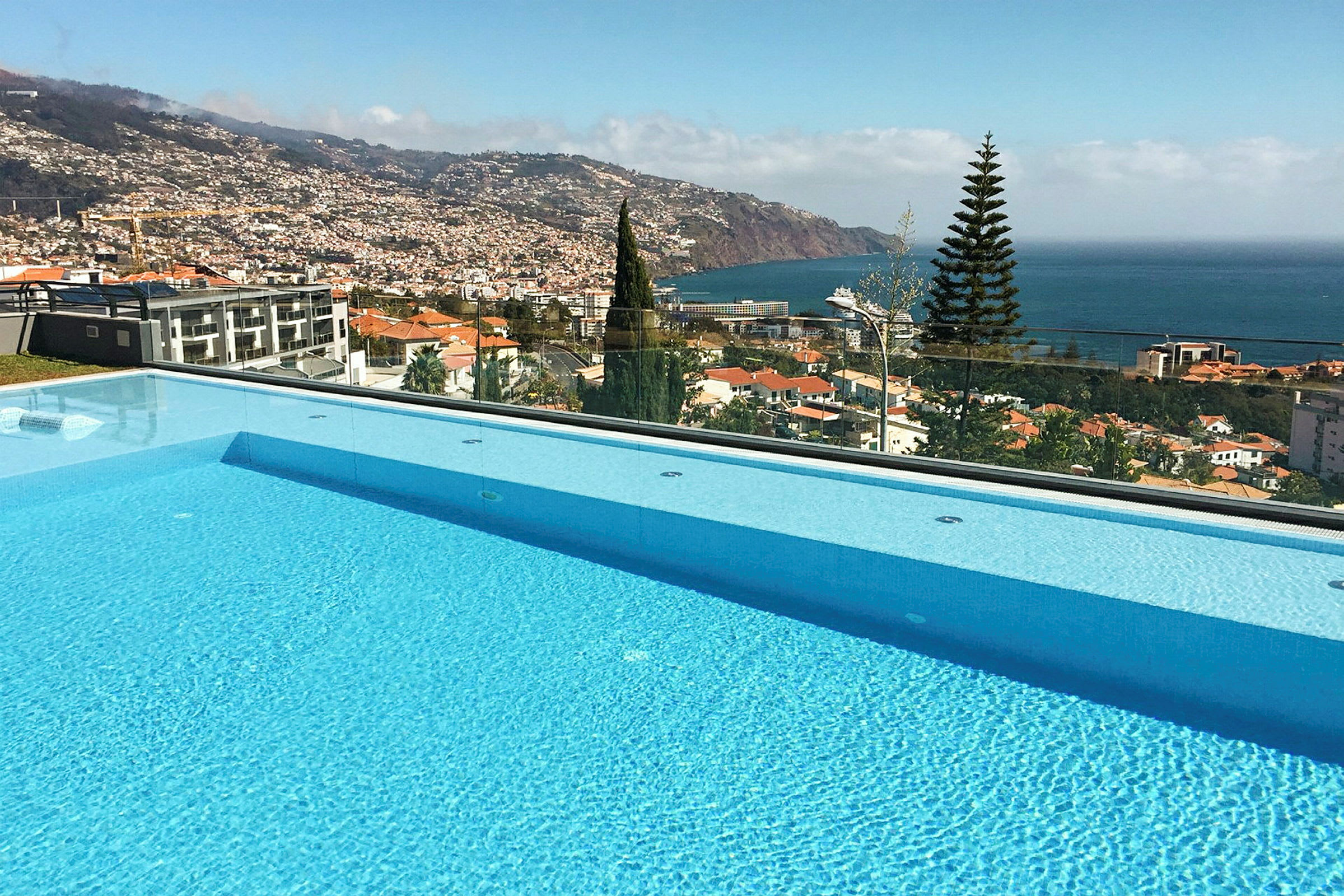 Utsikt över en pool med en vy av stadens hus samt havet och omgivande berg. (AI-genererad text)
