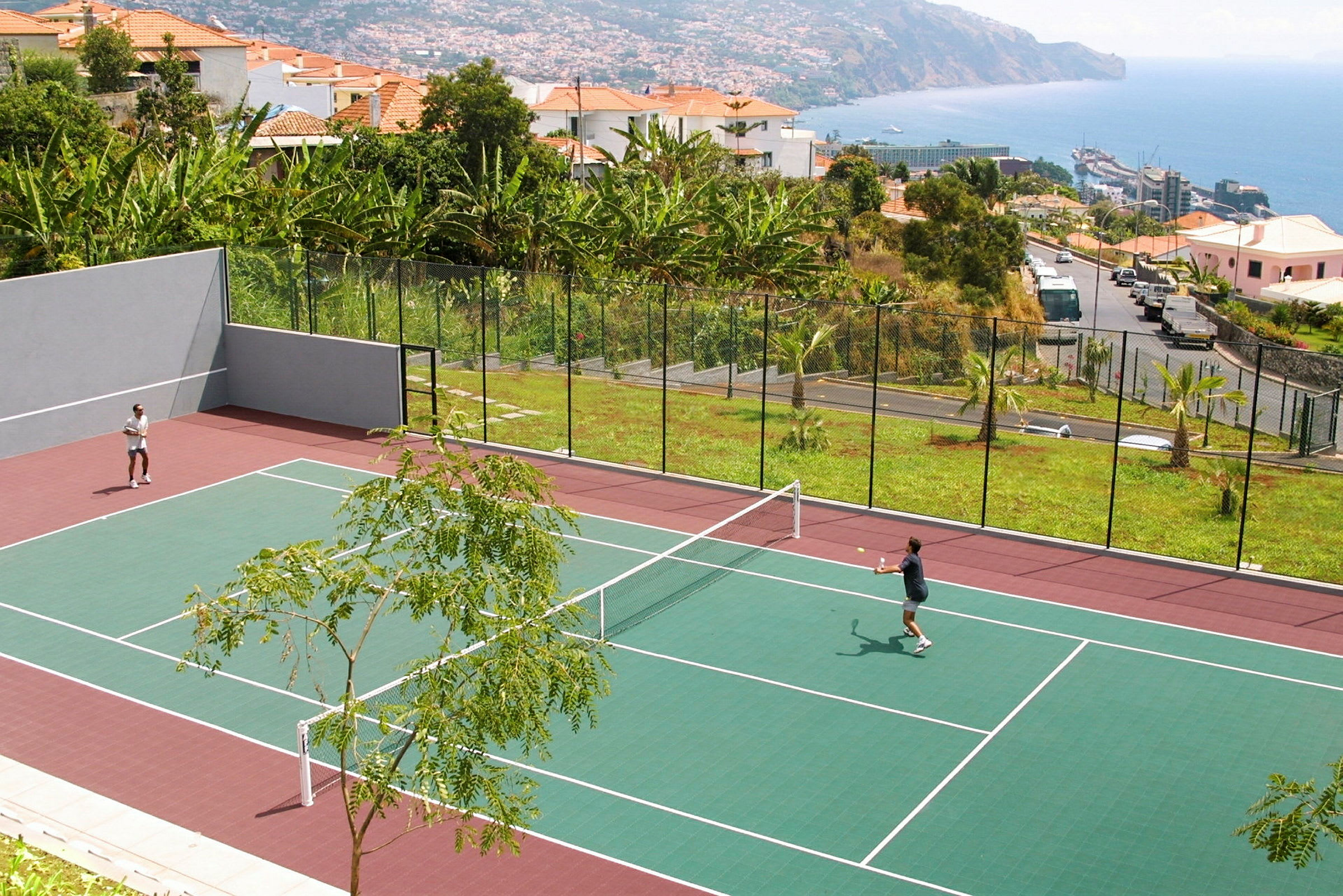 Två personer spelar tennis på en utomhusbana med omgivande grönska och havsutsikt. (AI-genererad text)