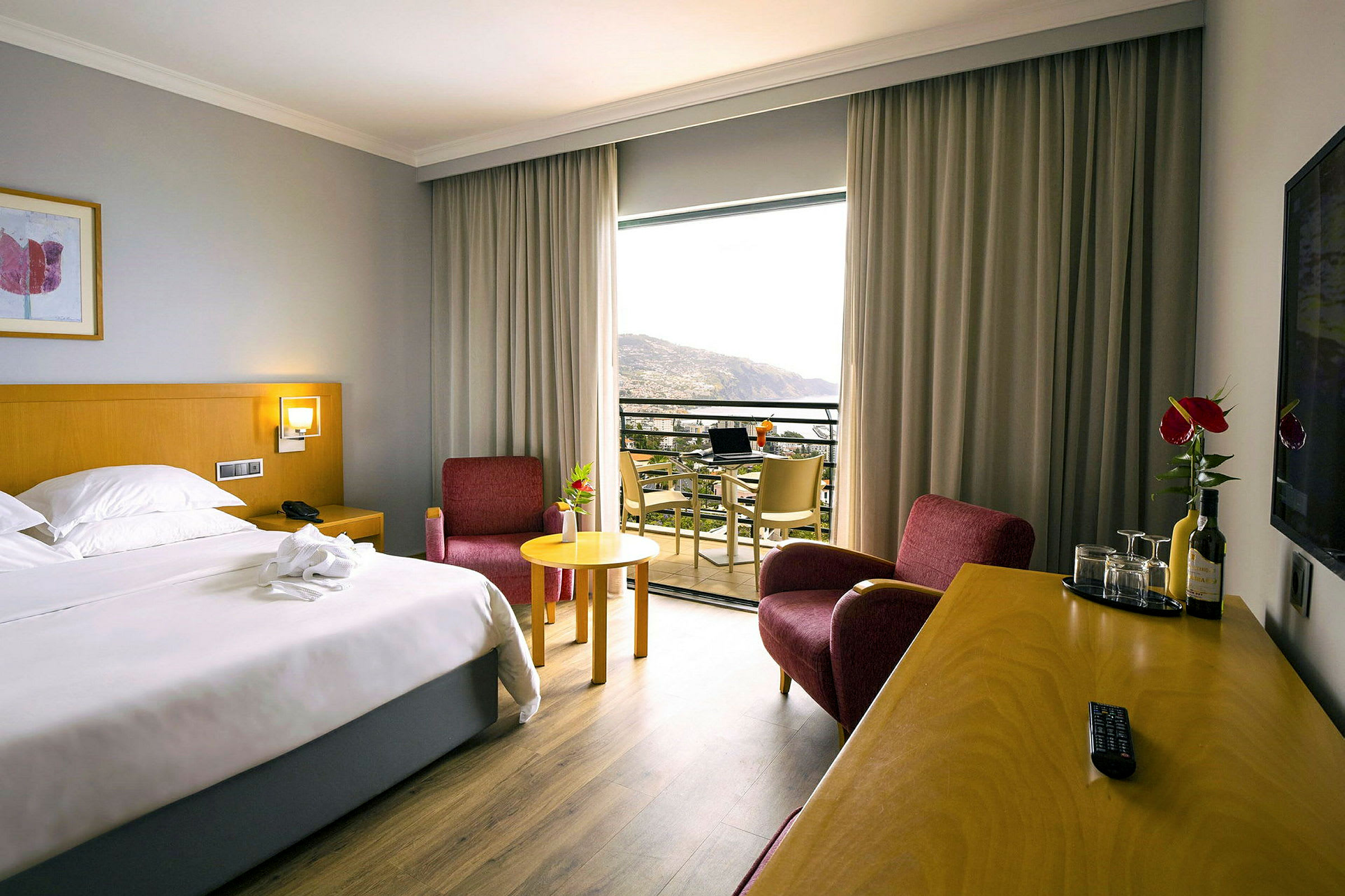 Hotellrum med en dubbelsäng, två röda fåtöljer, ett skrivbord med drycker och glas, och en balkong med utsikt. (AI-genererad text)