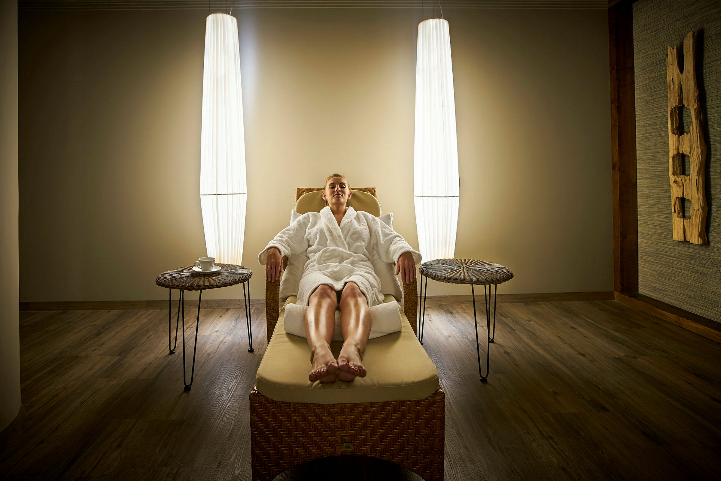 En person sitter avslappnad i en spa-lounge i en rottingstol, omgiven av mjuk belysning. (AI-genererad text)