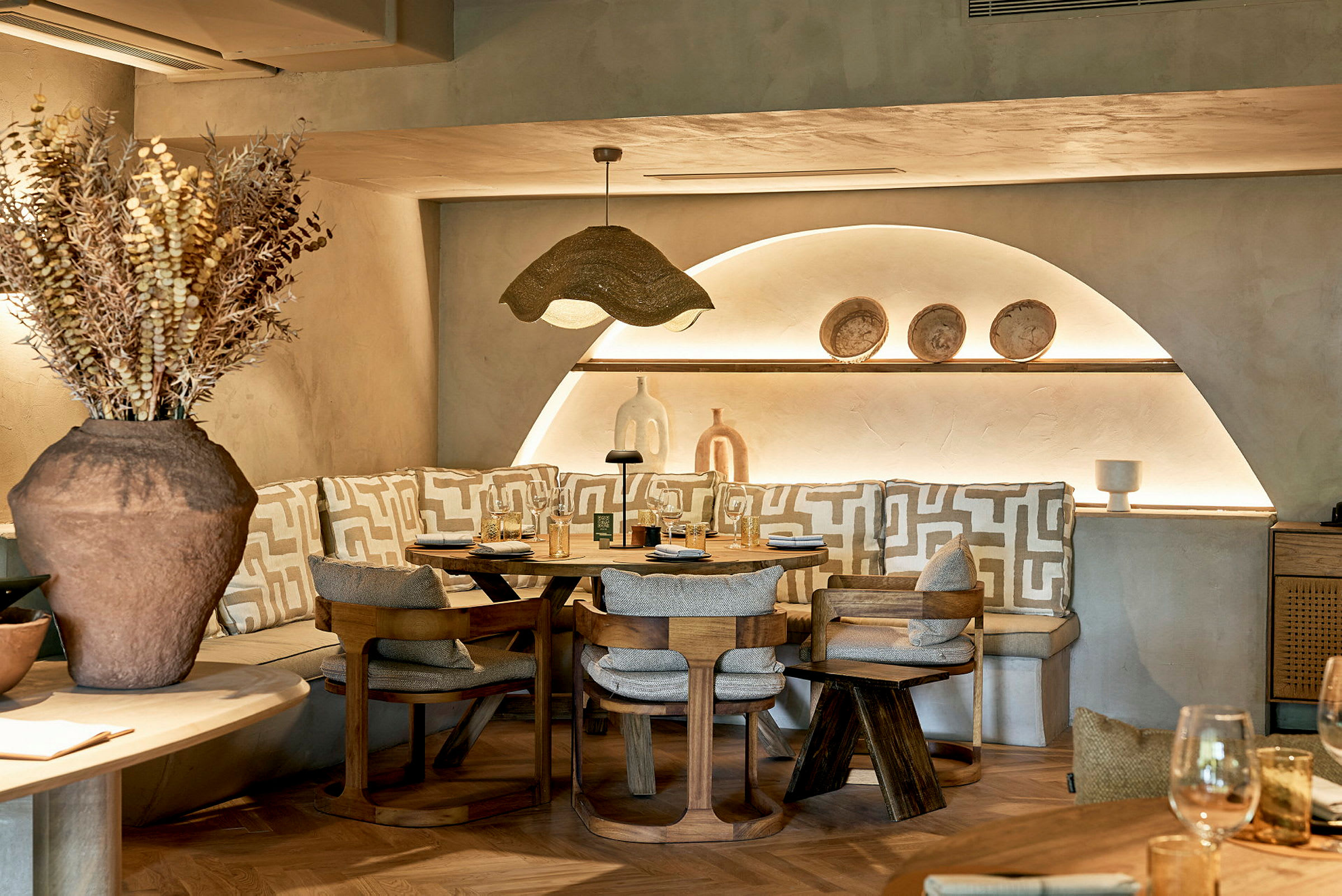 Ett mysigt restauranghörn med rundad sittplats, rustika trästolar och en dekorativ lampa ovanför bordet. På hyllan syns keramiska prydnadssaker. (AI-genererad text)