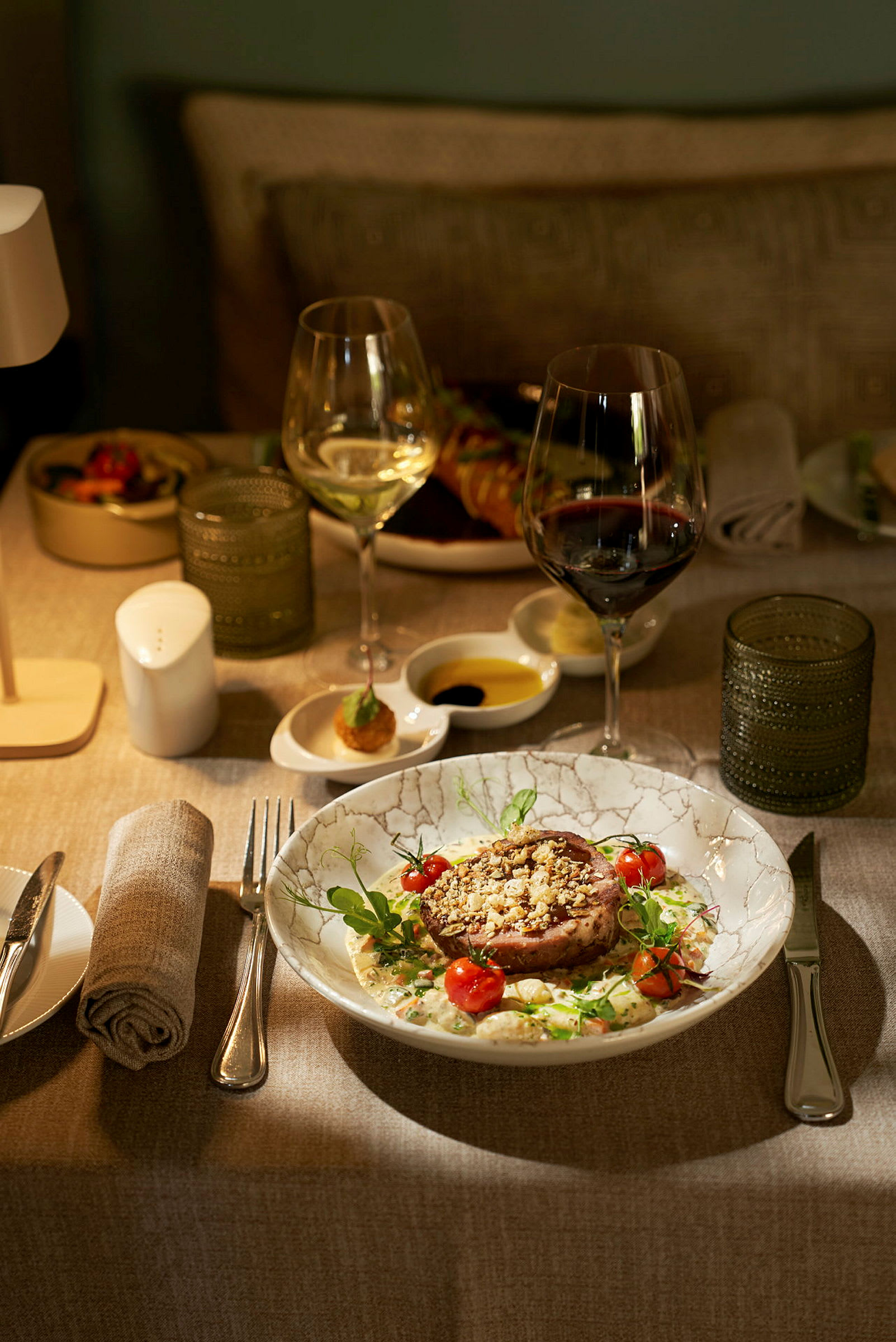 En tallrik med en fint upplagd måltid med kött och grönsaker, omgiven av vinglas, servett och andra bordstillbehör. (AI-genererad text)