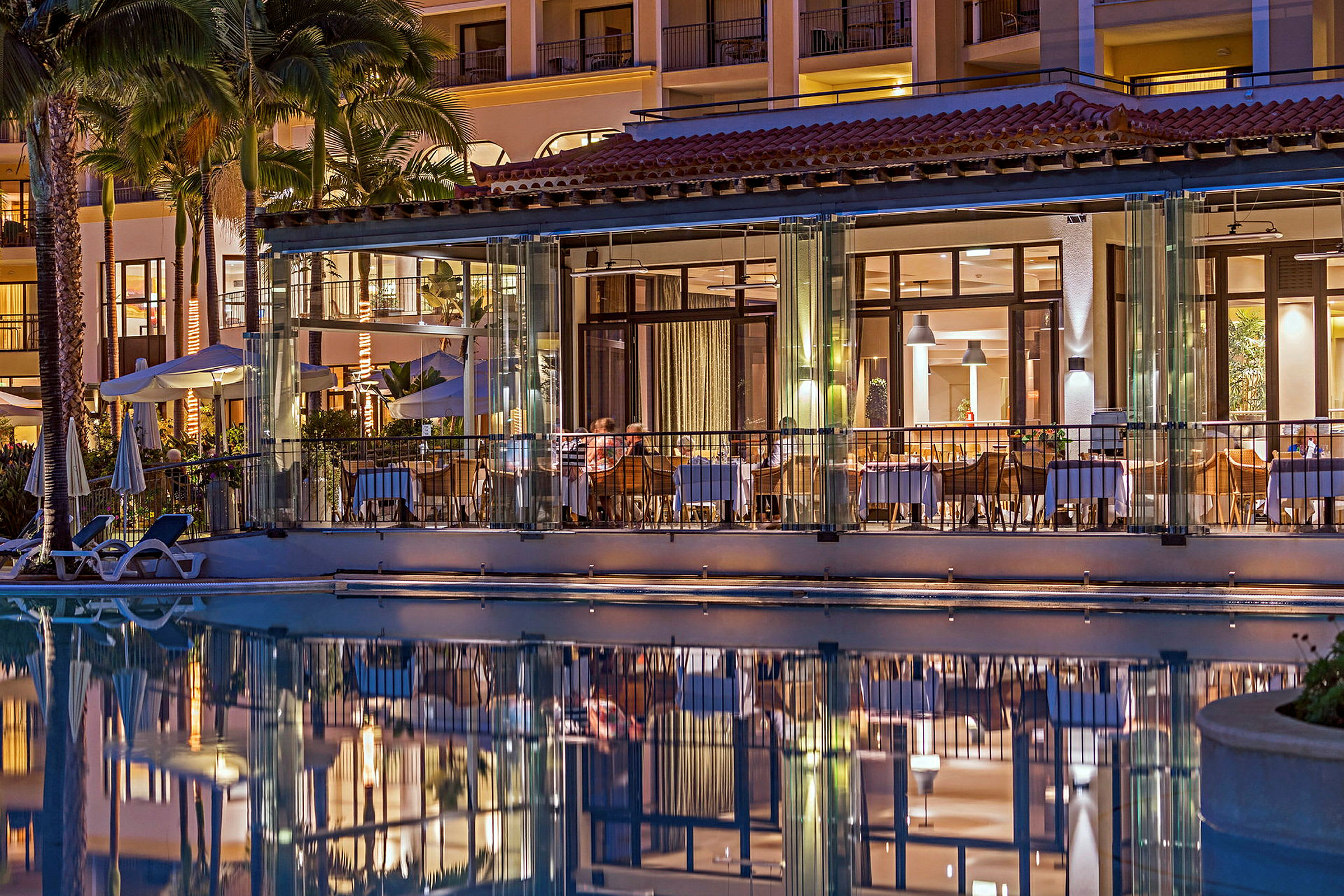 Utsikt över en upplyst hotellrestaurang med glasväggar och spegling i en närliggande pool. (AI-genererad text)