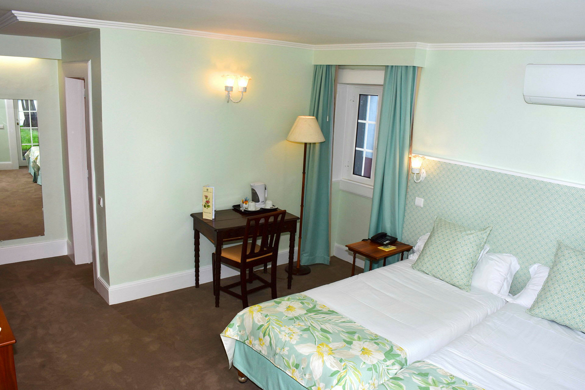 Ett hotellrum med två enkelsängar bäddade med vita lakan och gröna blommiga överkast. Rummet har ljusgröna väggar och ett skrivbord med en stol. (AI-genererad text)