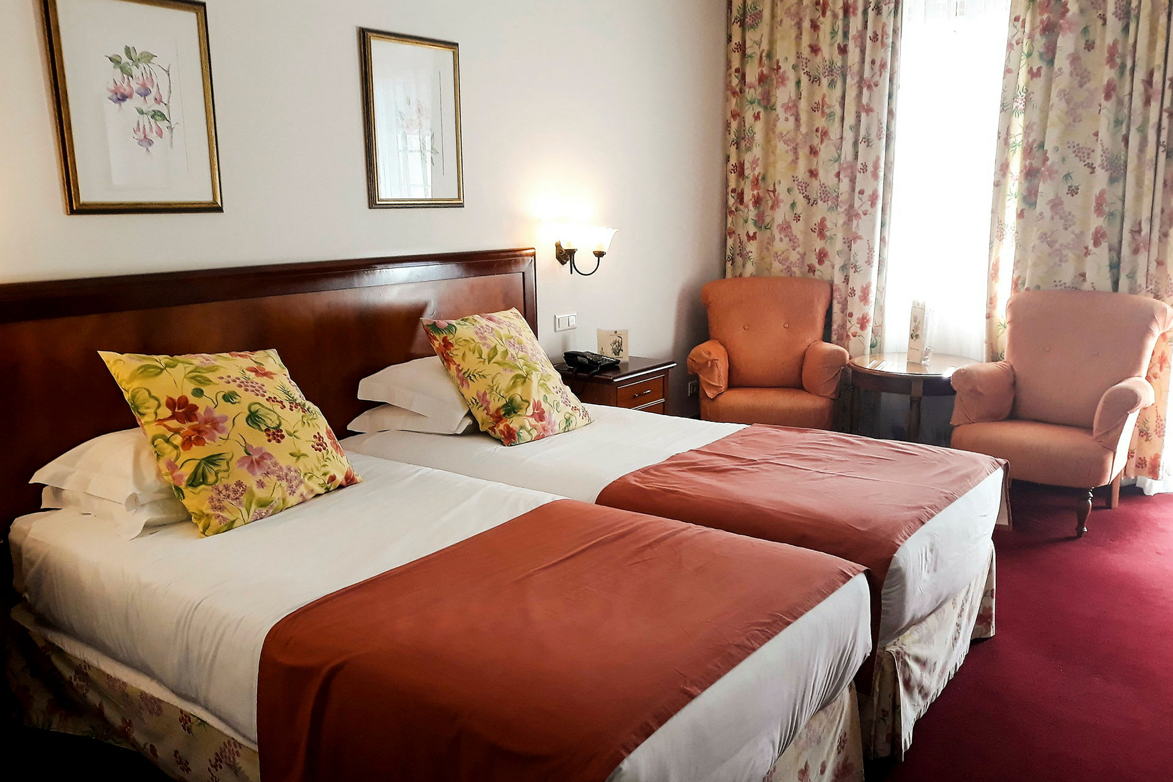 Ett hotellrum med två enkelsängar, dekorerade med blommiga kuddar och röda överkast. I rummet finns också två fåtöljer, ett litet bord och gardiner med blommönster. (AI-genererad text)