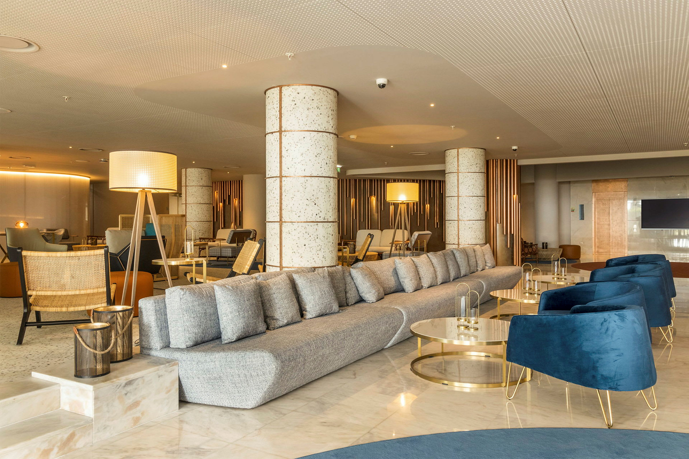 Ett modernt inrett hotellobbyområde med en lång grå soffa, blå fåtöljer och dekorativa belysningsarmaturer. (AI-genererad text)