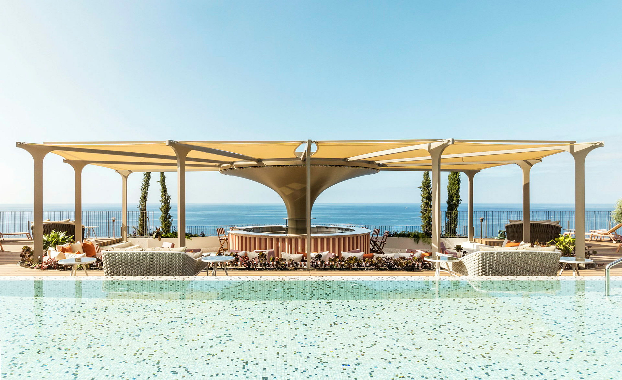 En pool med utsikt över havet och möblerade områden under skuggande tak. (AI-genererad text)