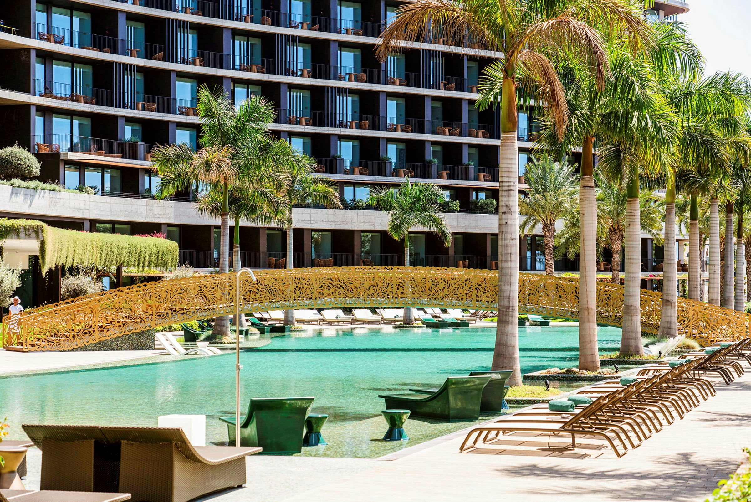 En pool med en dekorativ guldfärgad bro och omgivande palmer framför ett modernt hotell. (AI-genererad text)