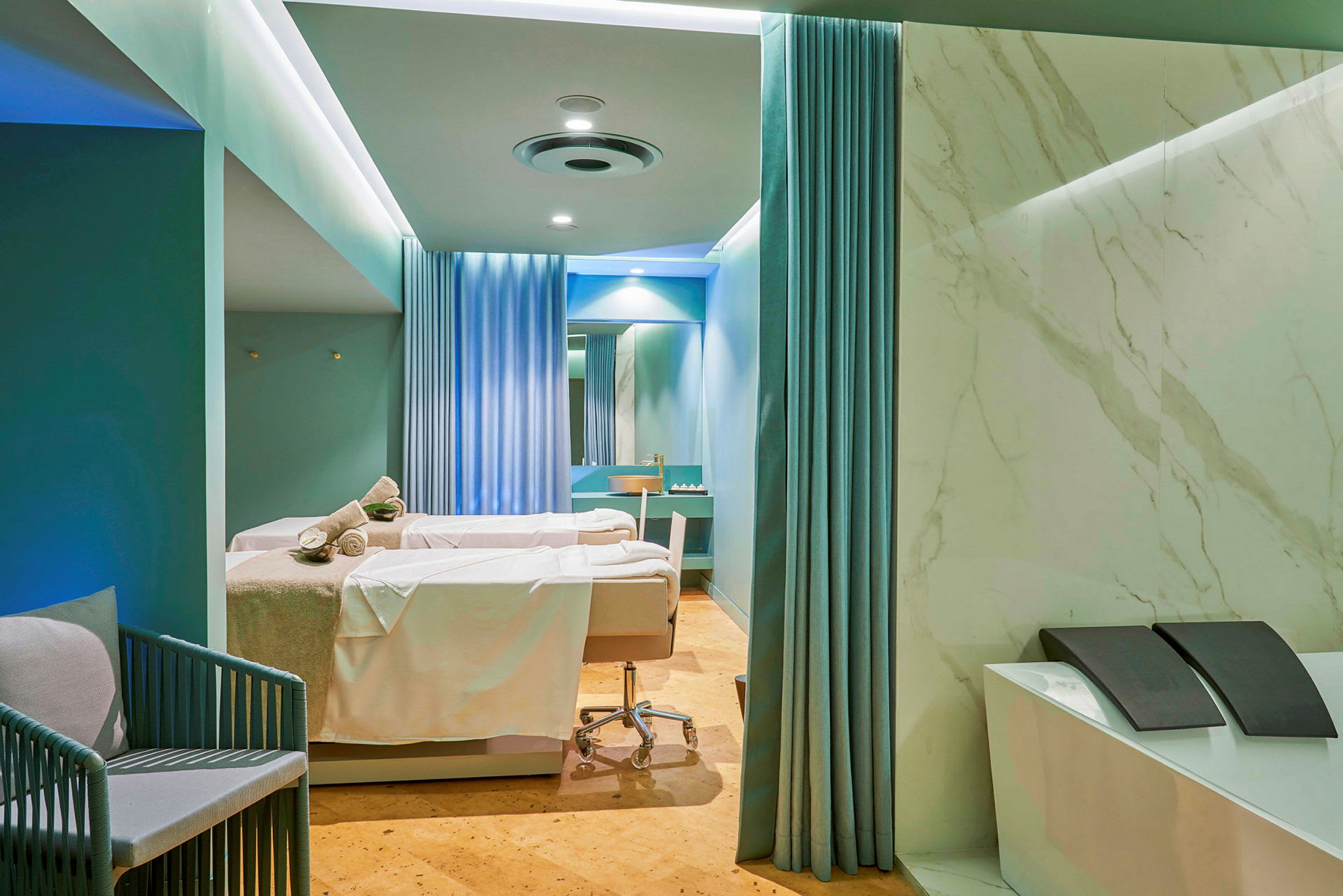 Ett spa- eller behandlingsrum med två behandlingssängar täckta av vita lakan och handdukar. Rummet är inrett med blå gardiner, marmorerade väggar och har en modern design. (AI-genererad text)