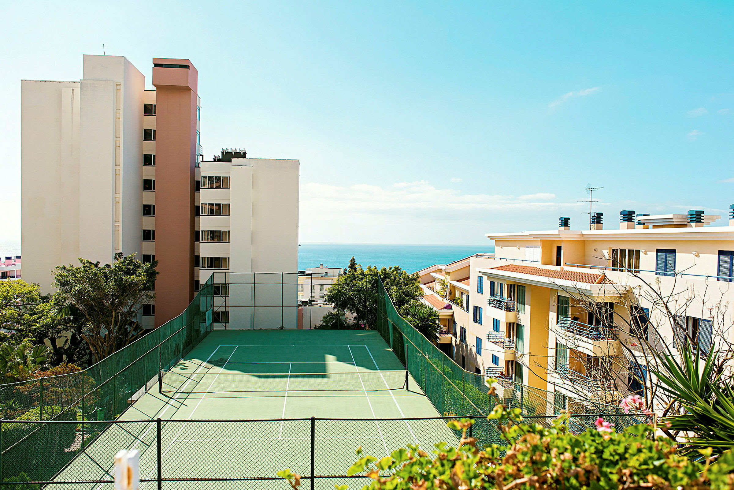Utsikt över tennisbana med höga byggnader och havet i bakgrunden. (AI-genererad text)