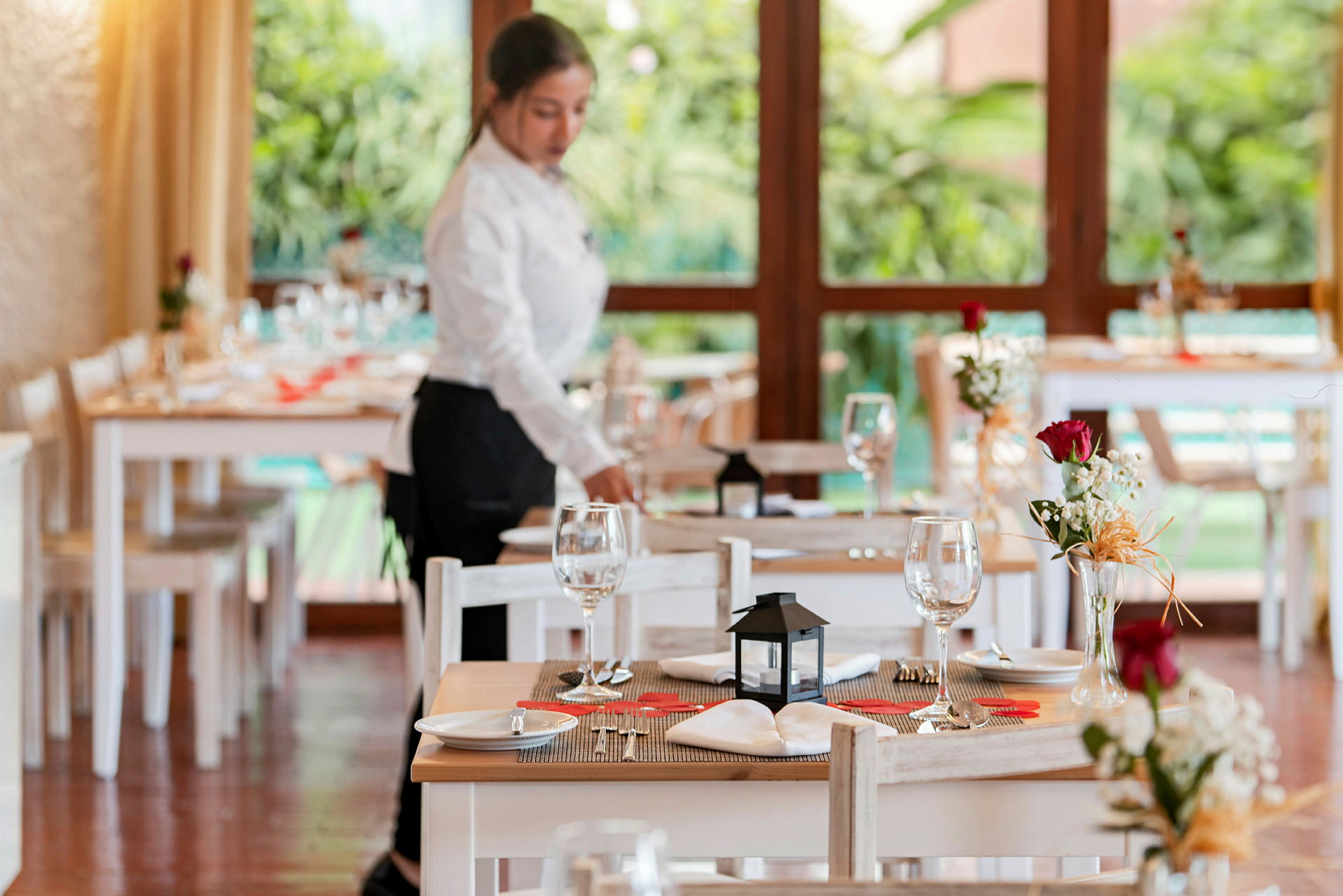 Ett dukat bord med bordsdekorationer och glas i en restaurang med stora fönster och gröna växter utanför. (AI-genererad text)