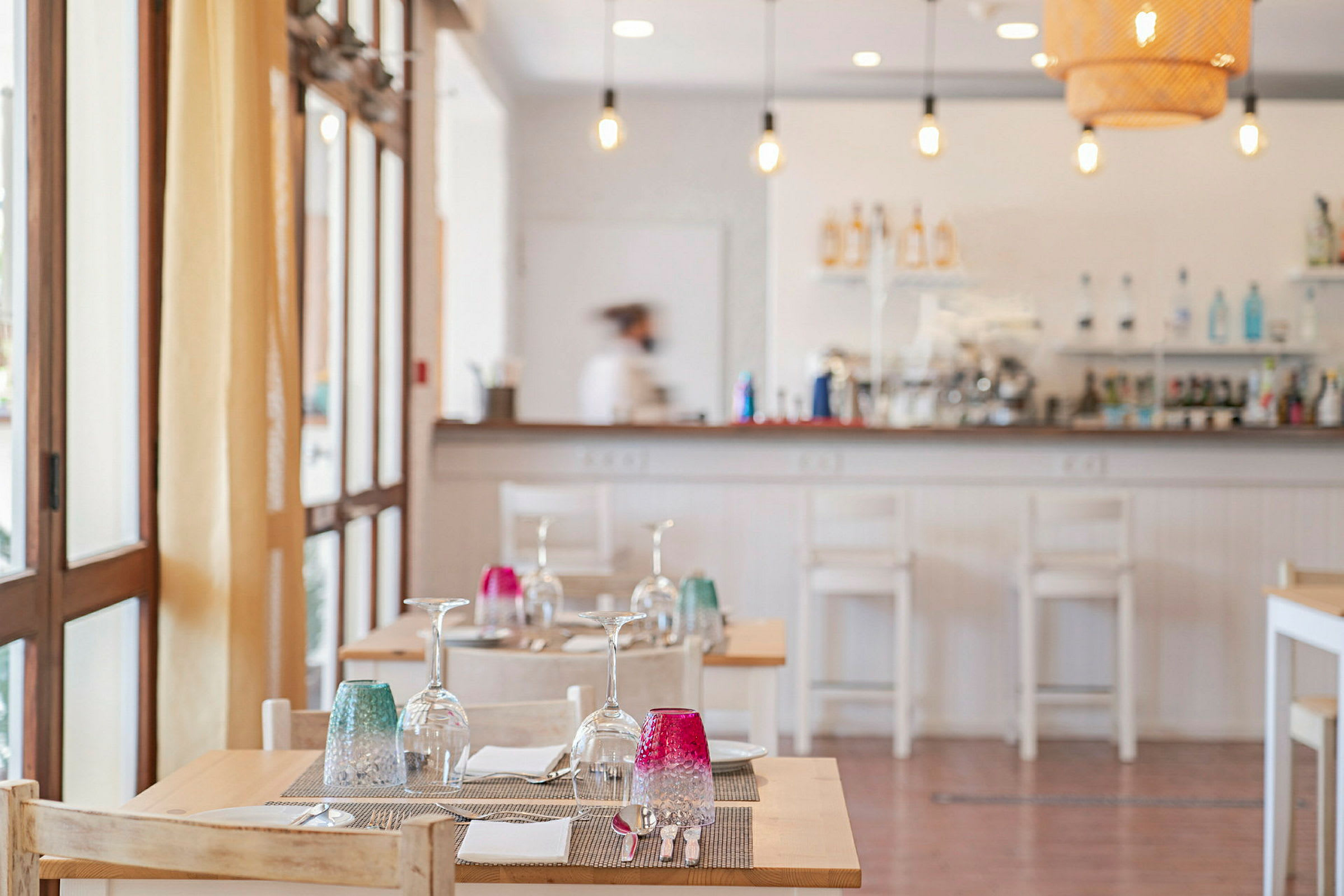 Interiör av en restaurang med dukade bord med glas och färgglada glas på. Servitör i bakgrunden vid bardisken. (AI-genererad text)