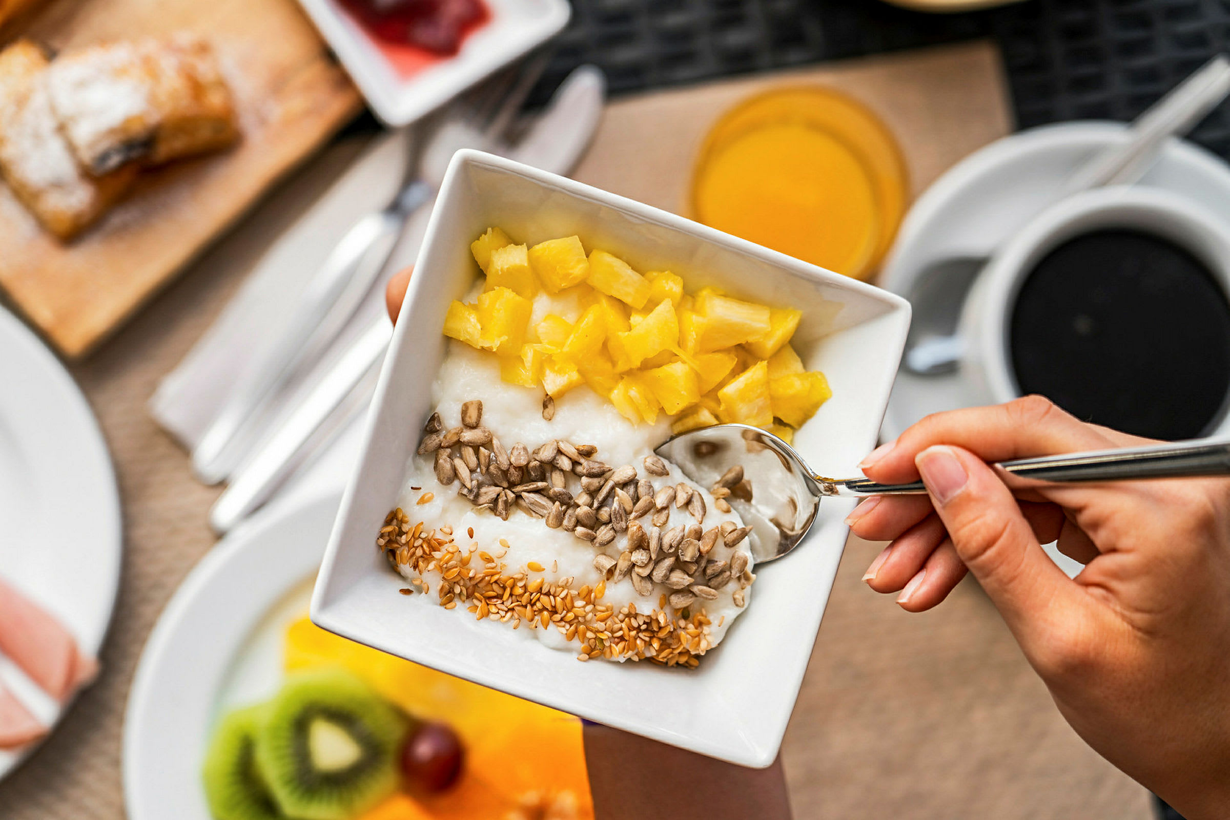 En hand håller en skål med yoghurt toppad med bitar av ananas, solrosfrön och sesamfrön. (AI-genererad text)