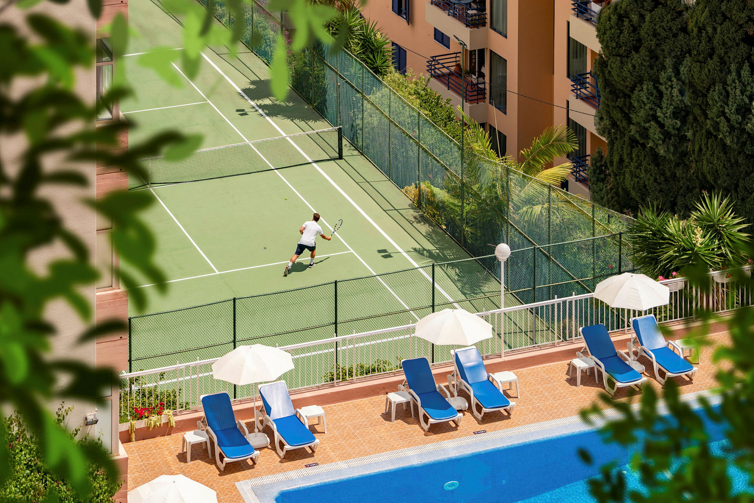 En person spelar tennis på en tennisplan nära en pool med solstolar och parasoller. (AI-genererad text)
