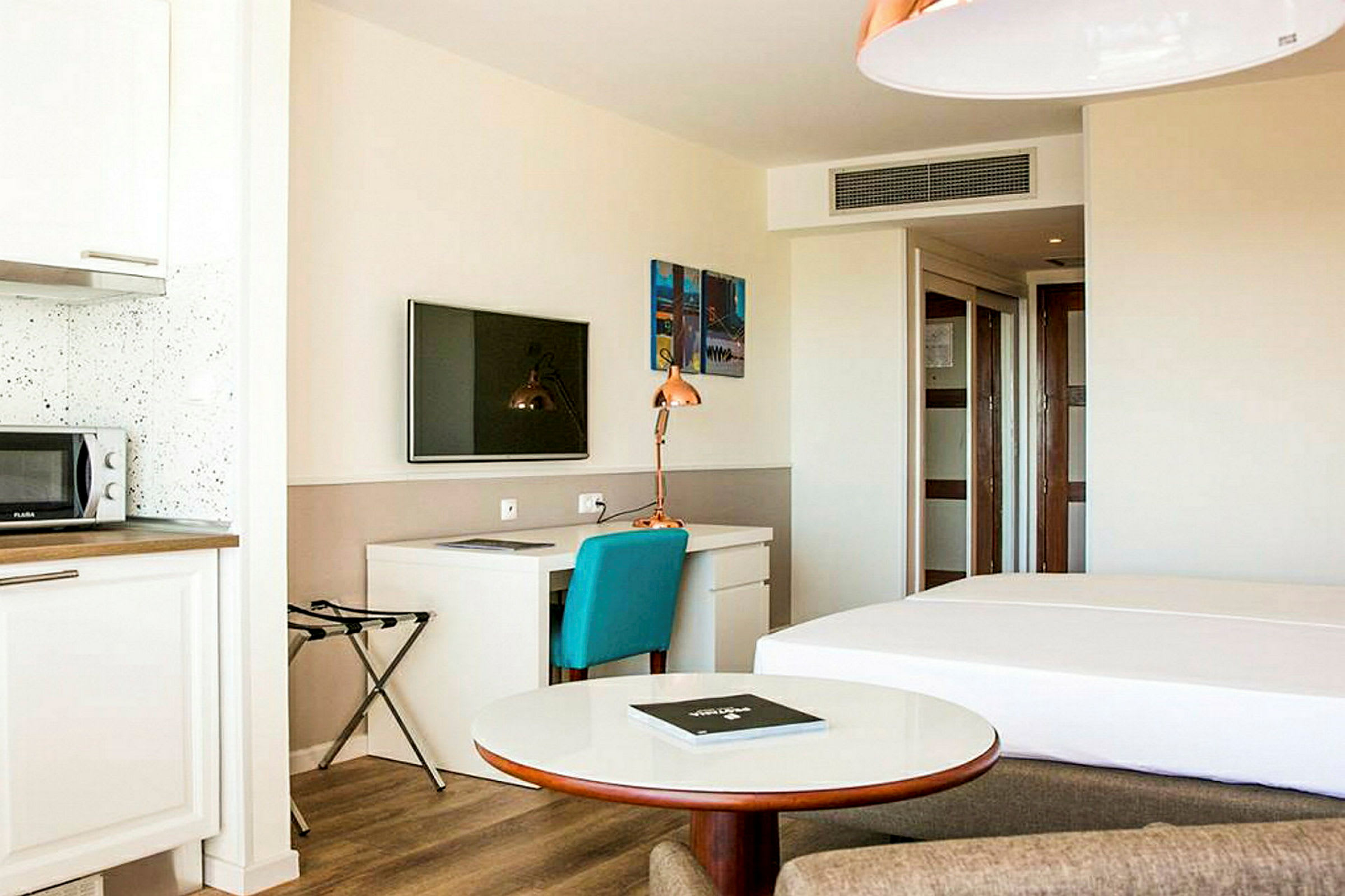 Ett ljust hotellrum med en säng, skrivbord, mikrovågsugn och en TV monterad på väggen. (AI-genererad text)