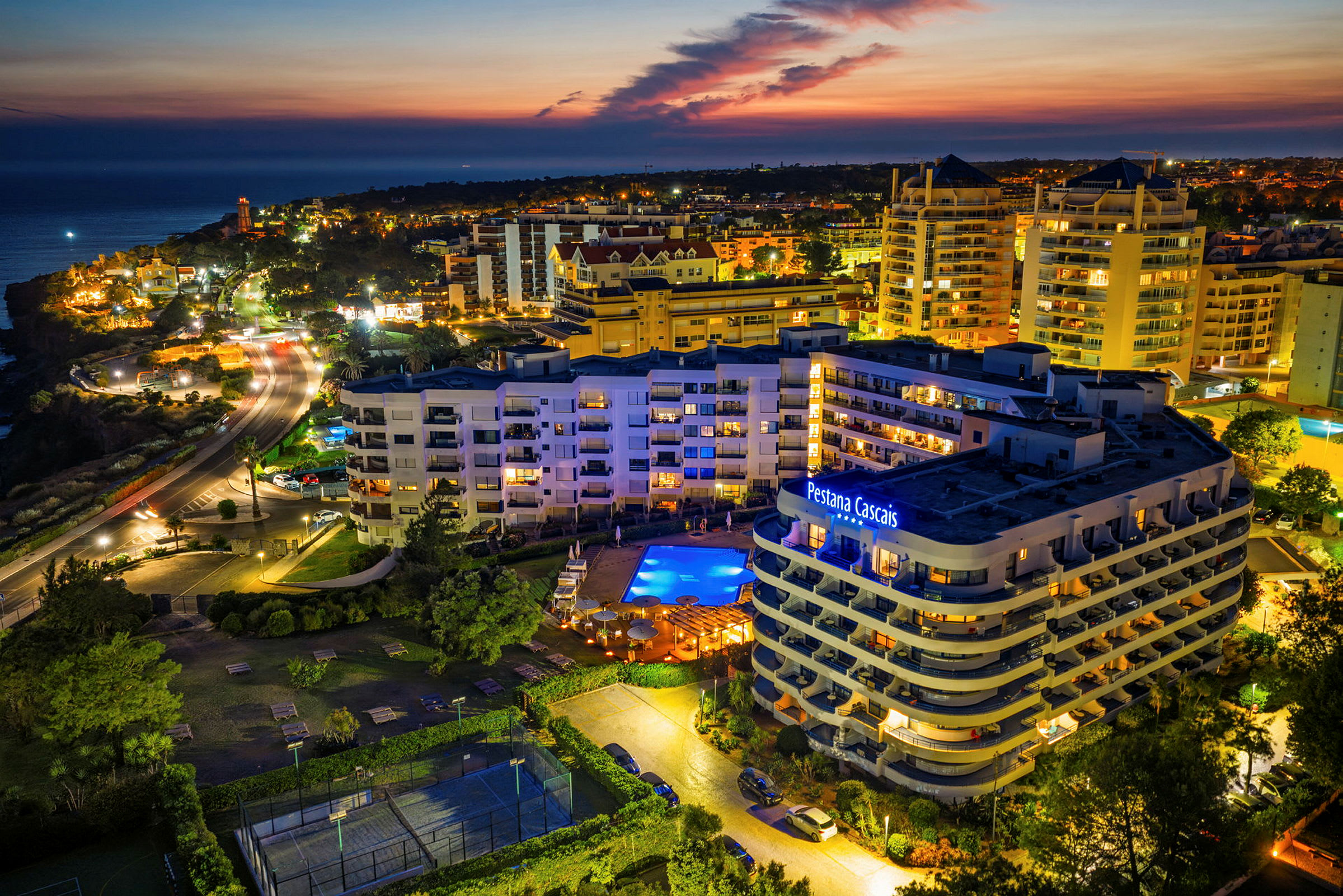 En vy över en upplyst hotellbyggnad och poolområde under skymningsljuset, med flerbostadshus och kustlinje i bakgrunden. (AI-genererad text)