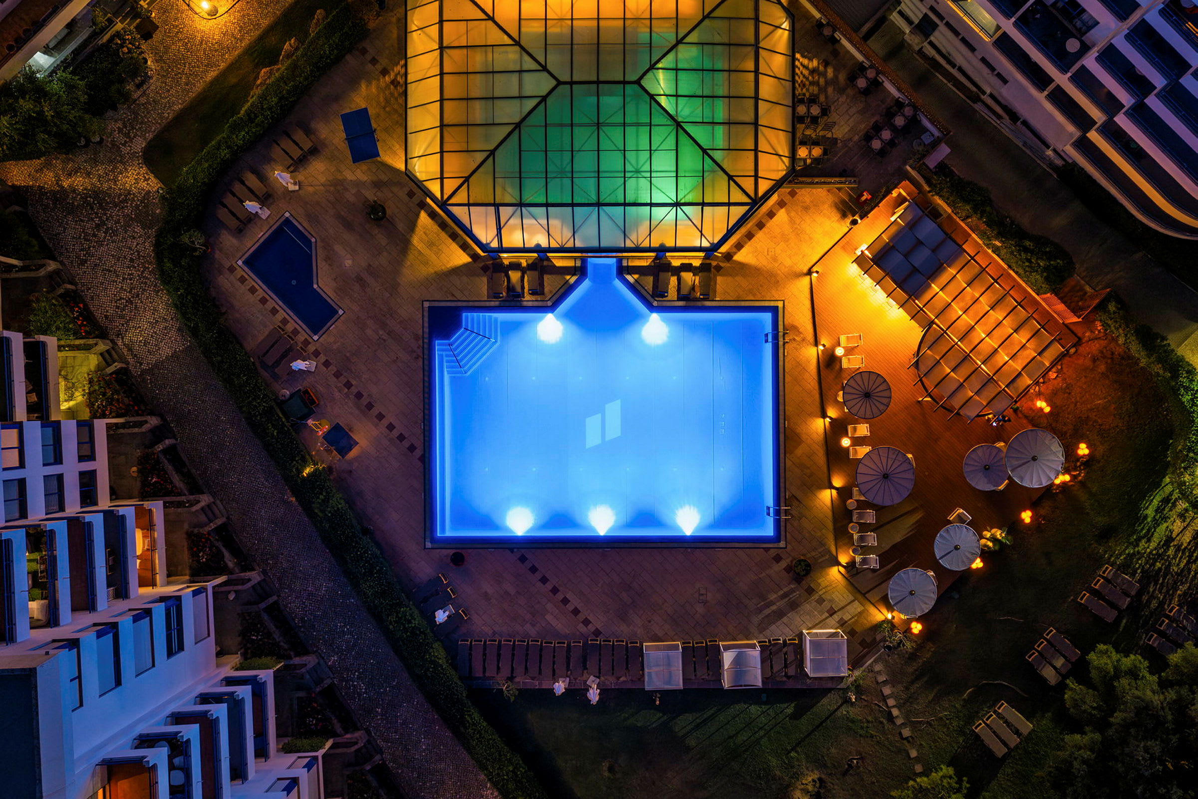 Flygfoto av en upplyst pool omgivande av byggnader och sittplatser. (AI-genererad text)